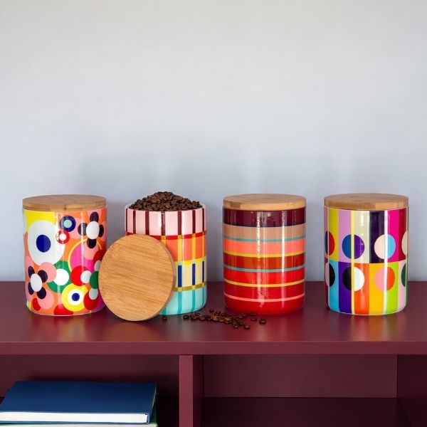 Quatre boîtes de conservation colorées avec couvercles en bois sur une étagère rouge foncé. La 2e à gauche est ouverte, avec des grains de café ; couvercle devant. Motifs modernes: fleurs, rayures, cercles jaunes, roses, bleus, rouges. Grains éparpillés.