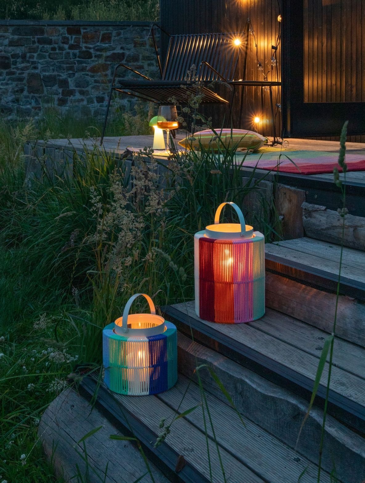 Zwei bunte Laternen mit warmem Licht stehen auf Holzstufen im hohen Gras. Die größere leuchtet Rot, Rosa und Grün, die kleinere Blau, Grün und Weiß. Im Hintergrund erhellen Lichterketten eine Terrasse mit Stuhl und Pilzlampe.