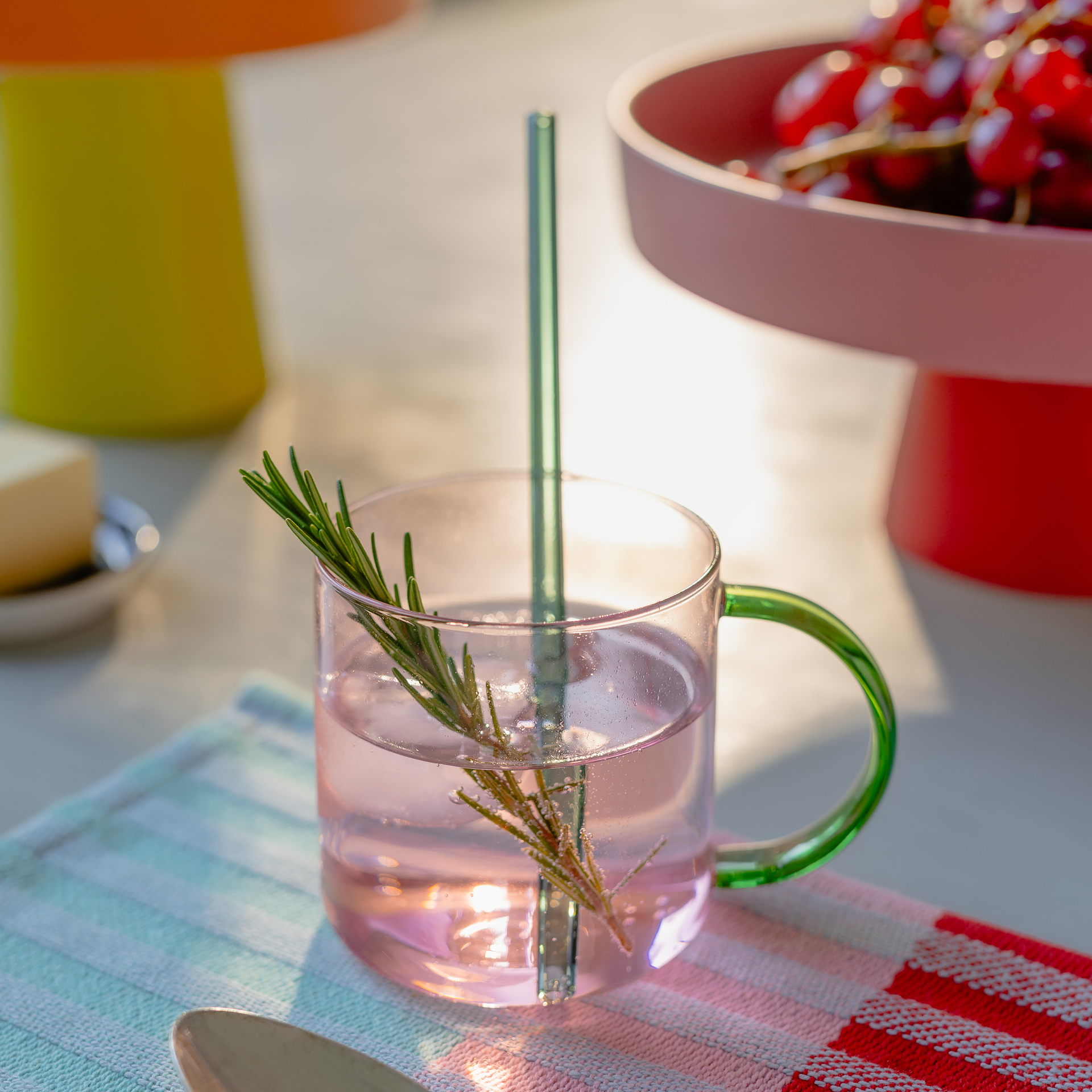 Ein rosefarbener Glasbecher 'Fred' mit grünem Henkel und Strohhalm, garniert mit Rosmarin, steht auf einer gestreiften Stoffserviette. Im Hintergrund Obstschale mit roten Trauben und eine gelbe Vase.