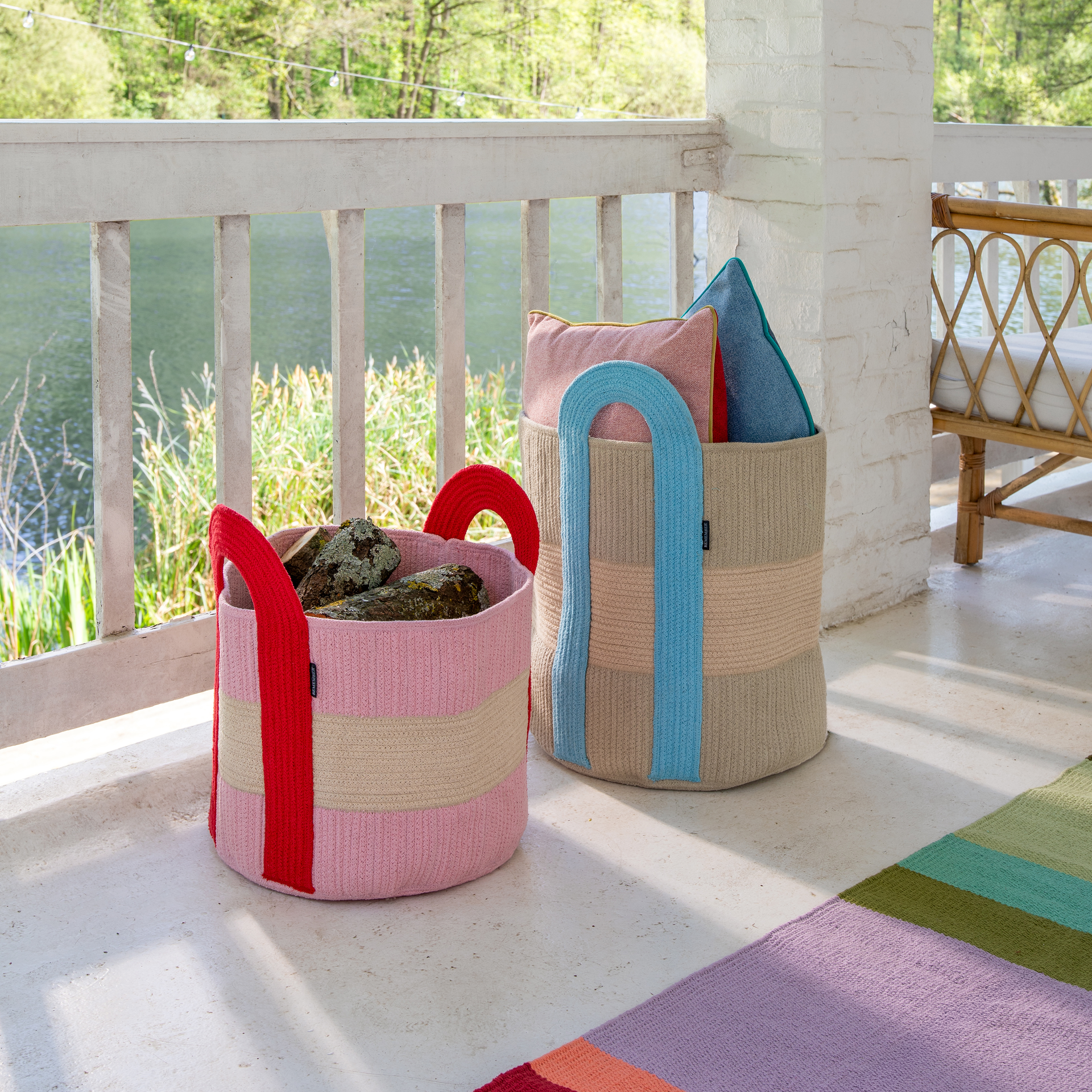 Zwei bunte, runde Aufbewahrungskörbe stehen auf einer Veranda am See. Der vordere Korb ist rosa-beige mit roten Griffen und Holzscheiten. Der hintere ist beige mit blauen Griffen und Kissen. Teils ist ein Teppich zu sehen.