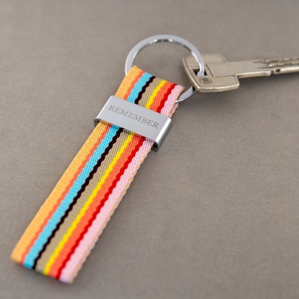 Porte-clés coloré sur fond gris : ruban en tissu à rayures verticales orange, turquoise, marron, jaune, rouge et rose. Il est maintenu par un curseur métallique argenté portant la gravure «REMEMBER » ; une clé argentée y est accrochée.