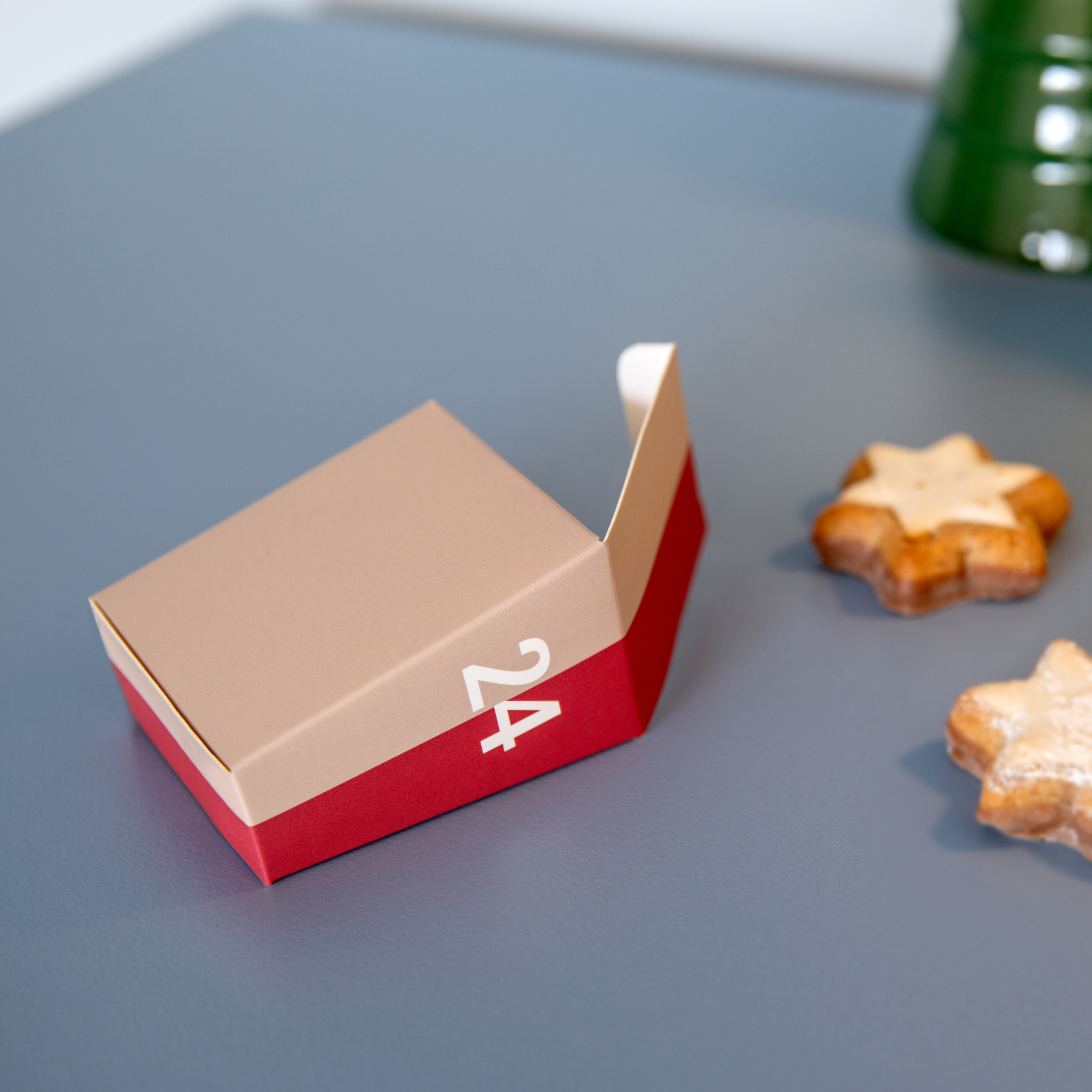 Boîte de calendrier de l'Avent rouge et beige ouverte, avec un « 24 » blanc sur fond gris. À droite, deux biscuits en forme d'étoile, légèrement saupoudrés de sucre glace et un peu flous, viennent rehausser l'ambiance festive.