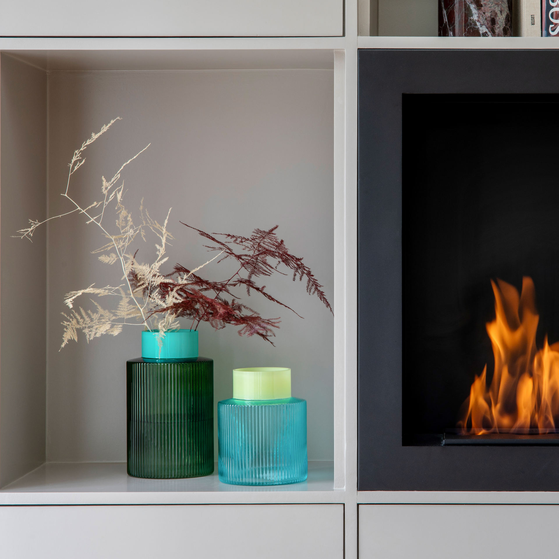 Im Regal stehen zwei Glasvasen, eine blaue und eine grüne Groove Vase, dekoriert mit getrockneten Zweigen, neben einem Kamin mit brennendem Feuer.