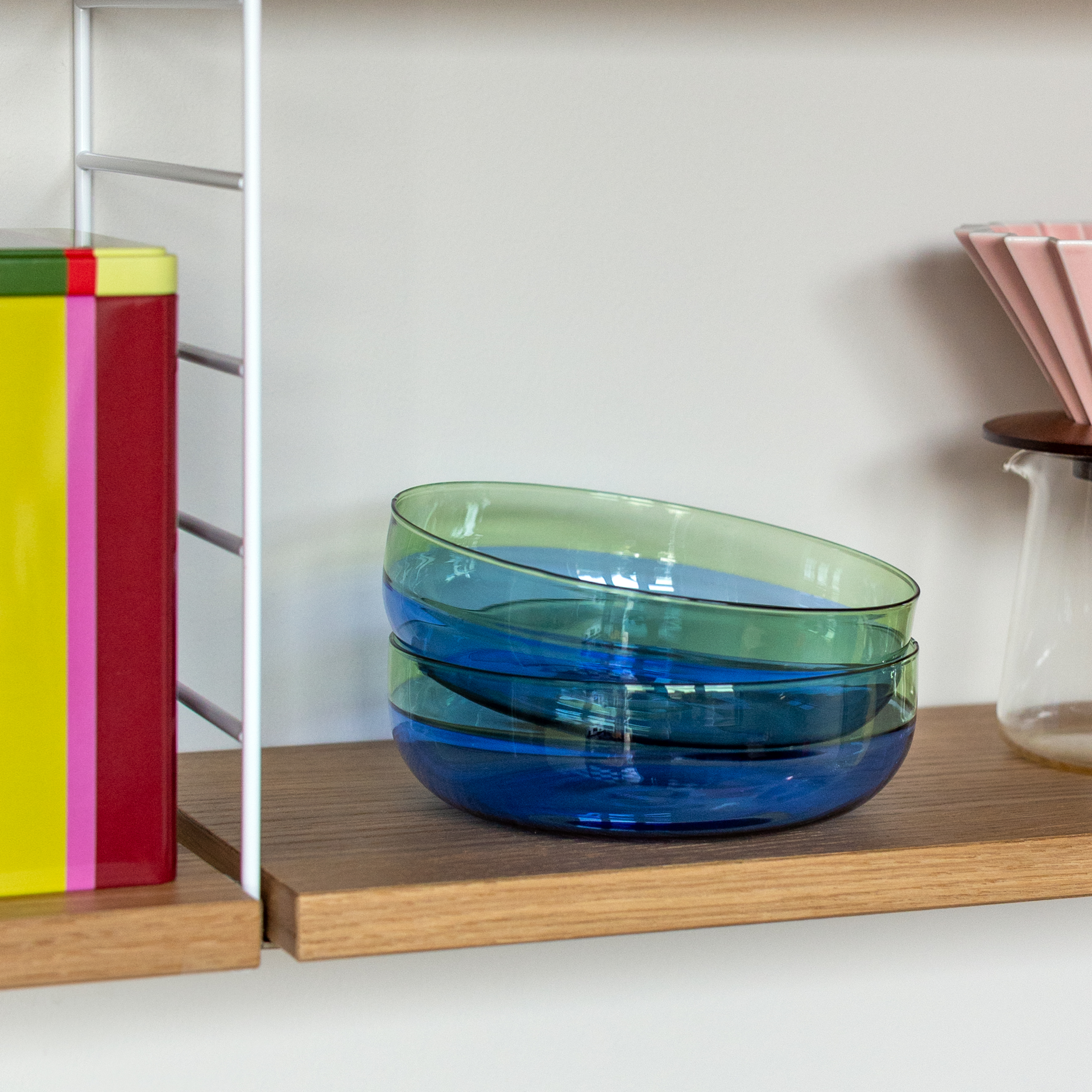 Auf einem Holzregal stehen zwei gestapelte Glasschalen, blau und grün. Links eine bunte Dose, rechts ein Kaffeefilter aus Glas mit rosa Papierfilter.