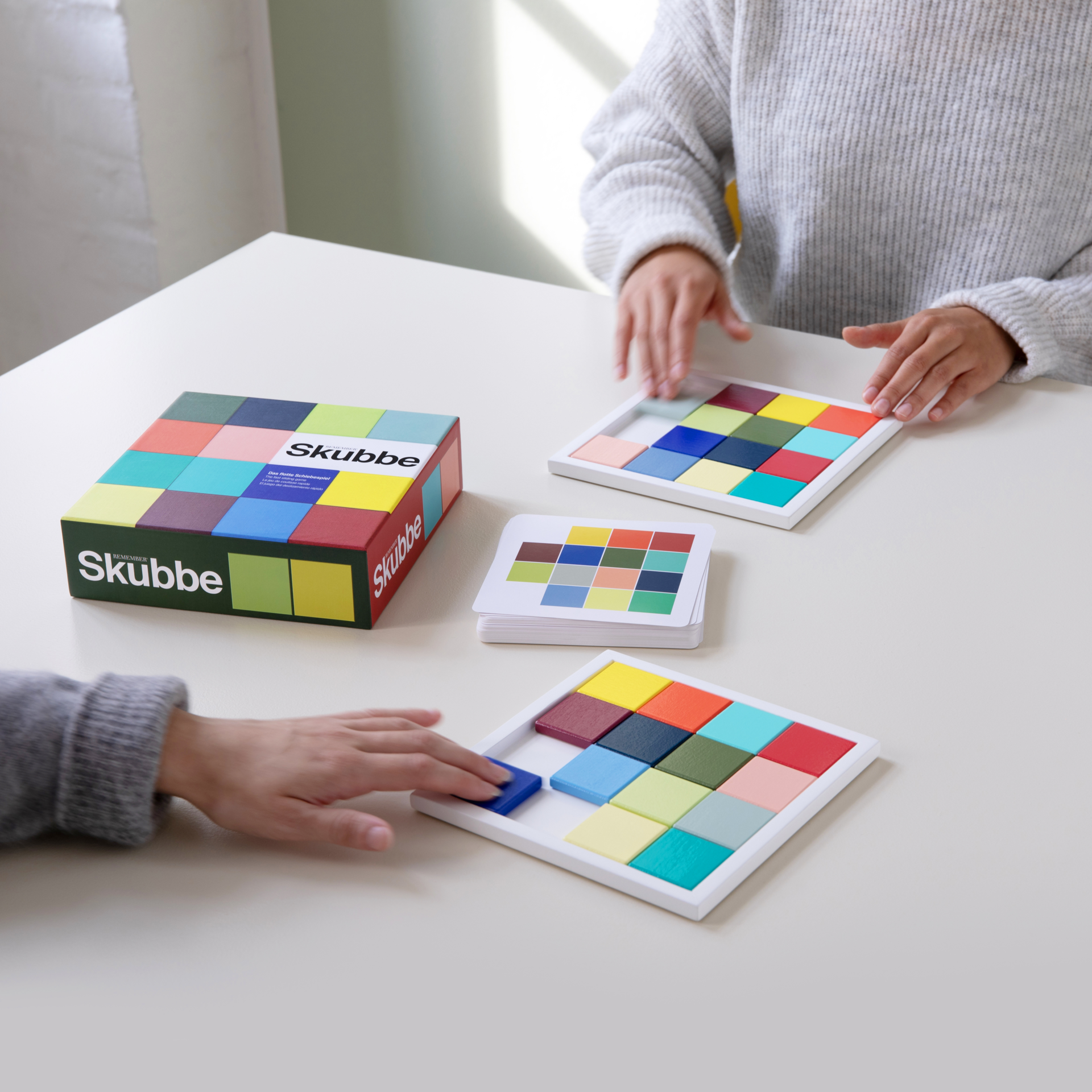 Zwei Personen spielen 'SKUBBE'. Holzspielsteine in Rahmen und Karten mit Farbmustern liegen auf einem Tisch. Die 'SKUBBE' Spielschachtel ist zu sehen.