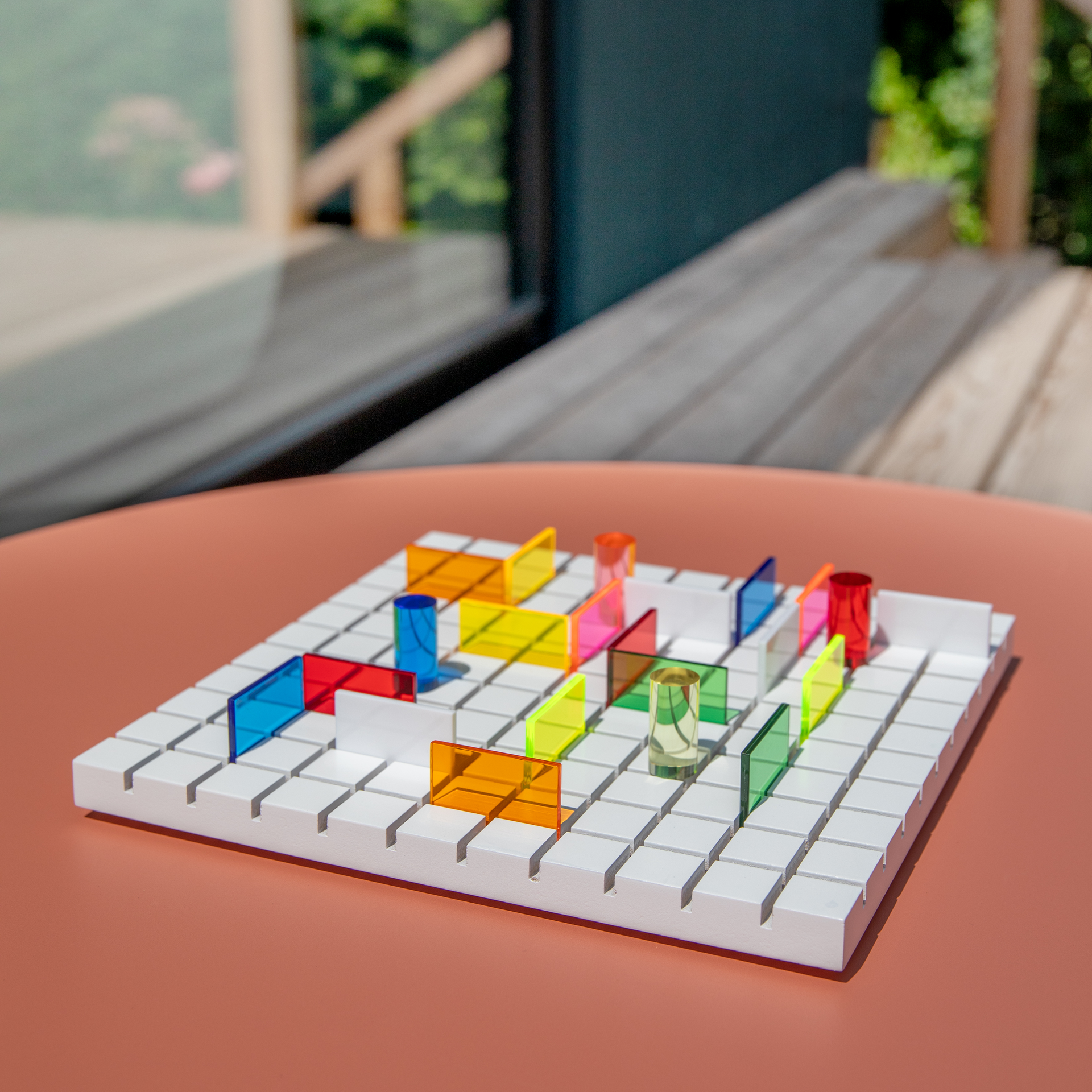 Weißes, quadratisches Spielbrett mit Gittermuster, darauf viele transparente, farbige Acrylelemente: rote, blaue, gelbe, grüne, orange Blöcke und zylindrische Säulen wie ein abstraktes Labyrinth. Terrakottatisch, unscharfe Natur im Hintergrund.