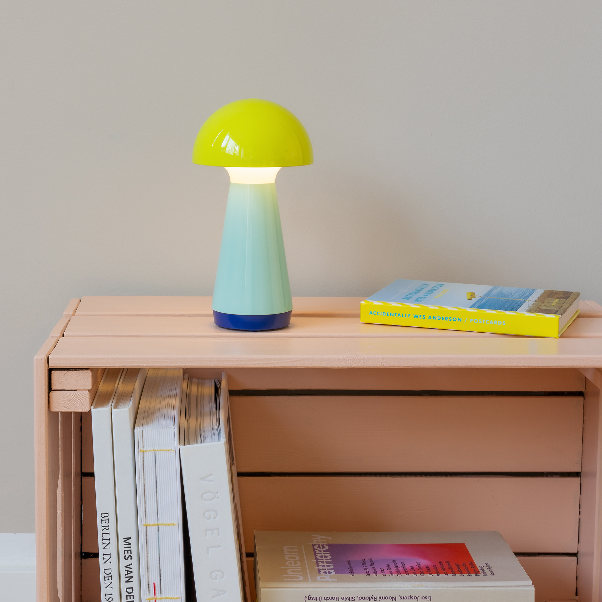 Lampe in Pilzform mit gelbem Schirm und blau-türkisfarbenem Stiel auf einem rosafarbenen Regal. Daneben liegen Bücher und eine Postkartensammlung von Wes Anderson.