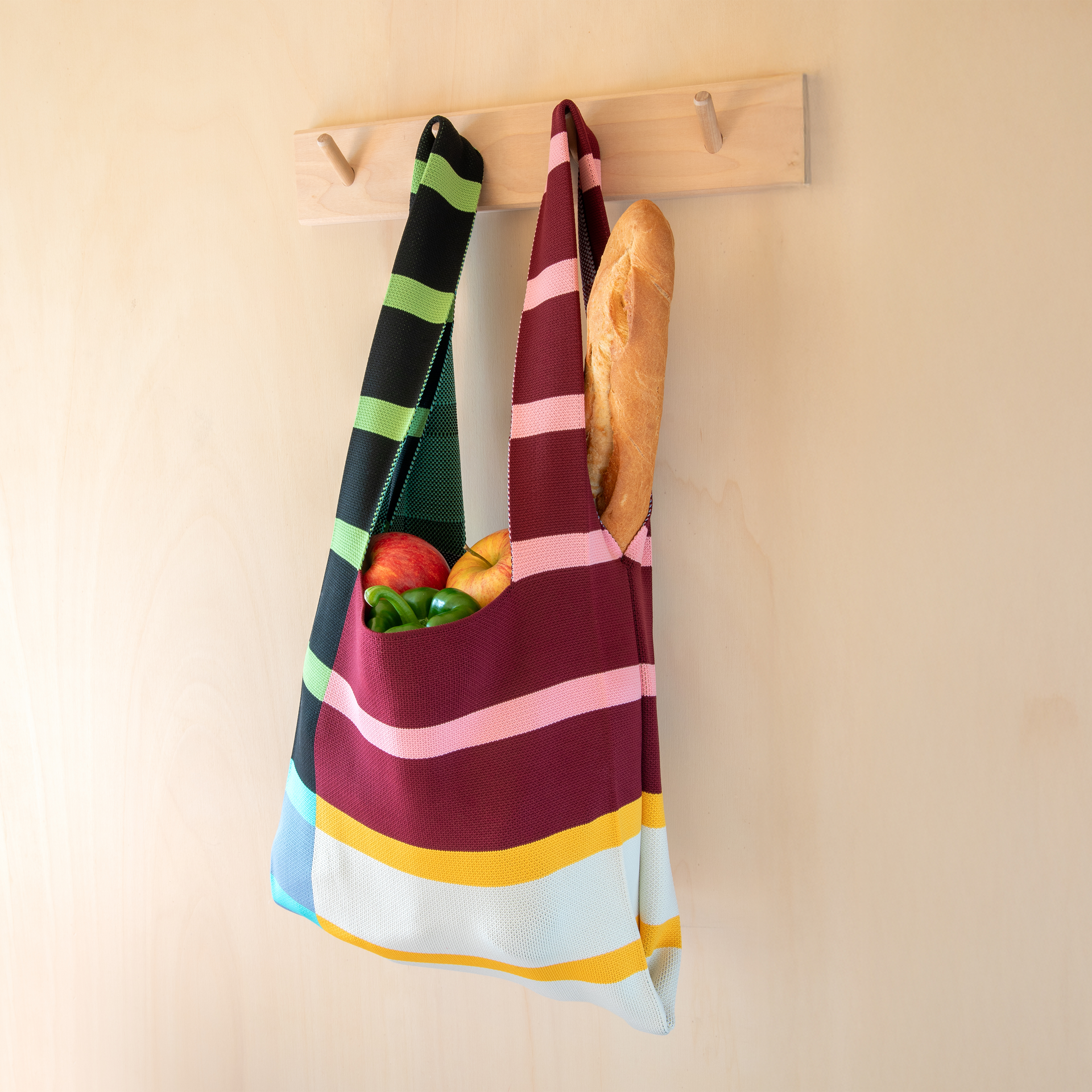Ein farbenfrohes, gestreiftes Strick-Accessoire mit zwei Henkeln auf weißem Hintergrund. Das Produkt weist ein Muster aus horizontalen Streifen in Grün, Schwarz, Rosa, Bordeaux, Gelb und Beige auf.