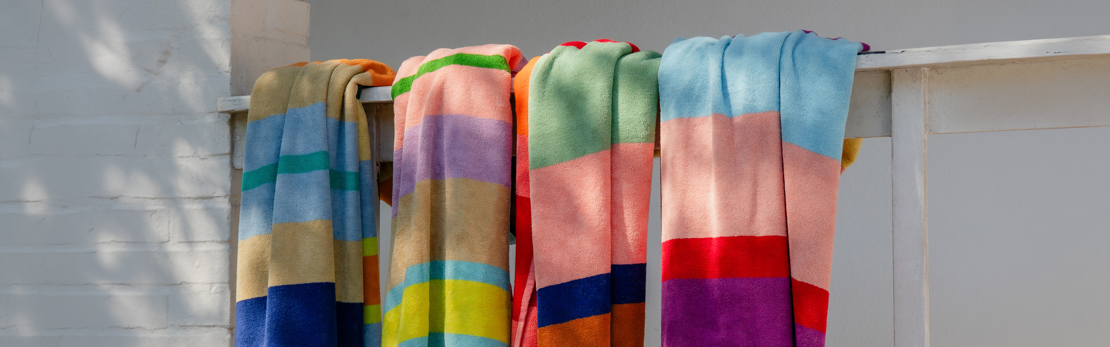Vier weiche, gestreifte Badetücher in leuchtenden Farben wie Blau, Gelb, Grün, Rosa, Lila und Orange hängen ordentlich über einer weißen Holzstange. Dahinter eine helle, leicht schattige Ziegelwand.
