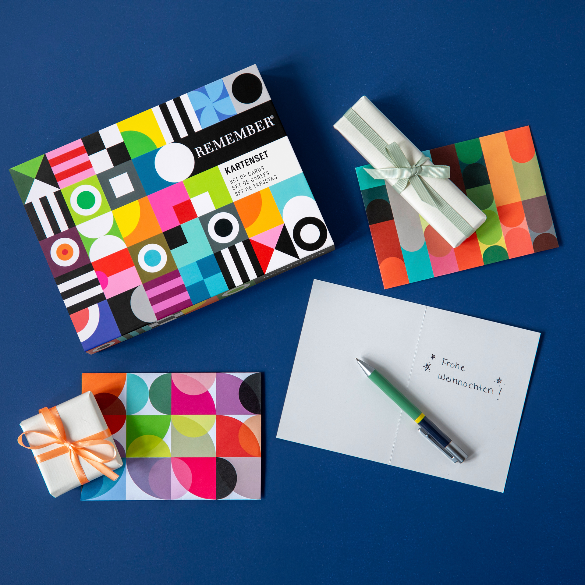 Karton mit dem Kartenset Remember, geöffneter Brief mit Stift und Geschenke auf blauem Hintergrund. Geometrische Muster und Farben.