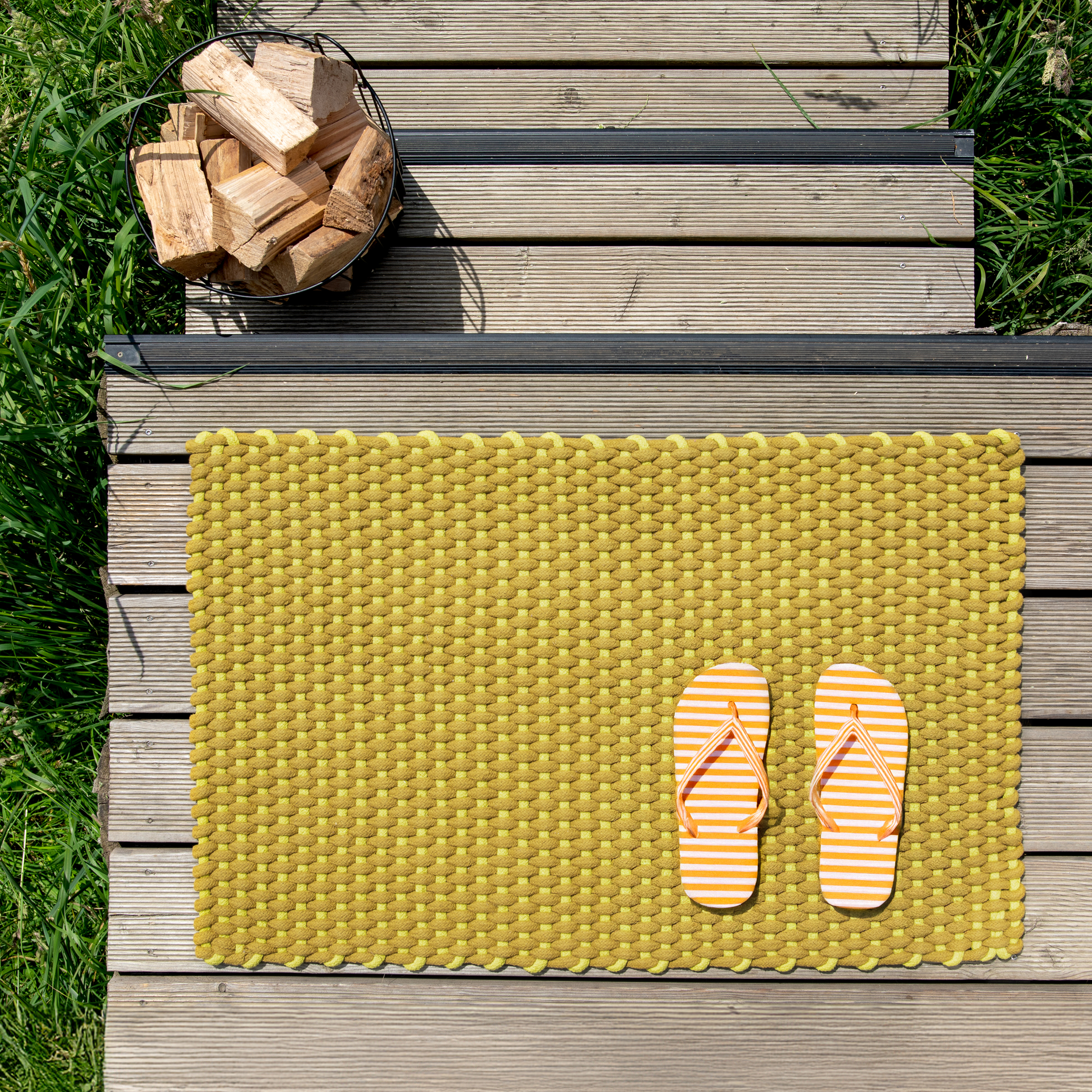 Handgewebte, lime-farbene Fußmatte mit orange-weiß gestreiften Flip-Flops auf einer Holzterrasse, neben einem Korb mit Holzscheiten und Gras.