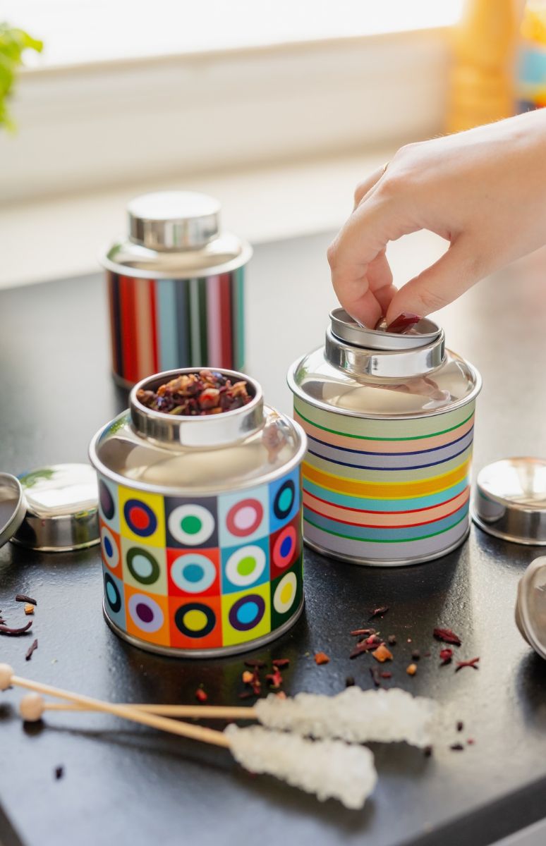 Eine Hand öffnet eine bunte, gestreifte Teedose auf dunkler Oberfläche. Daneben steht eine Dose mit bunten Kreisen auf Quadraten, gefüllt mit losem Tee. Im Hintergrund eine dritte Dose. Teeblätter und zwei Zuckerstäbchen liegen daneben.