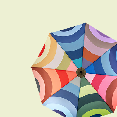 Draufsicht eines bunten Sonnenschirms vor hellgrünem Hintergrund. Das Design besteht aus Farbkreisen in Segmenten, die sich von der Mitte aus spiralförmig nach außen winden. Farben umfassen Rot, Blau, Grün und Rosa.