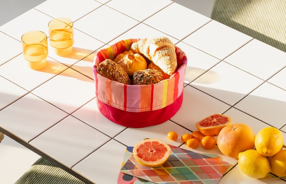 Auf einem weiß gefliesten Tisch steht ein farbenfroher Brotkorb mit frischen Brötchen und Croissant. Daneben zwei gelbe Trinkgläser und Zitrusfrüchte: halbierte Grapefruits, Zitronen und kleine Kumquats im Sonnenlicht.