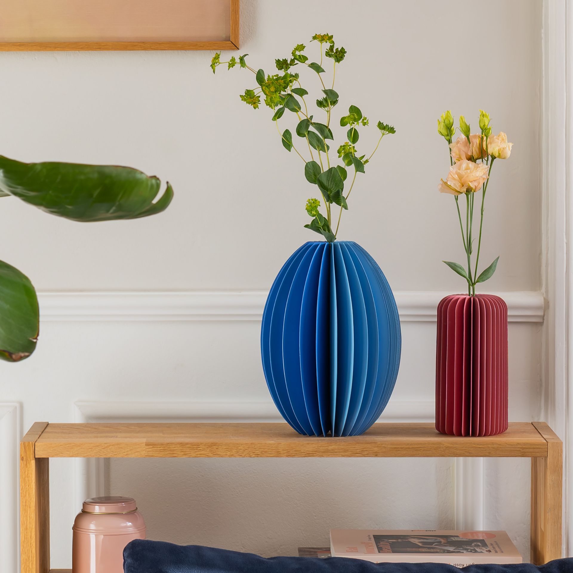 Auf einem Holzregal stehen eine blaue, kugelförmige und eine rote, zylindrische Vase, beide mit Blumen darin. Links ist eine grüne Pflanze sichtbar.