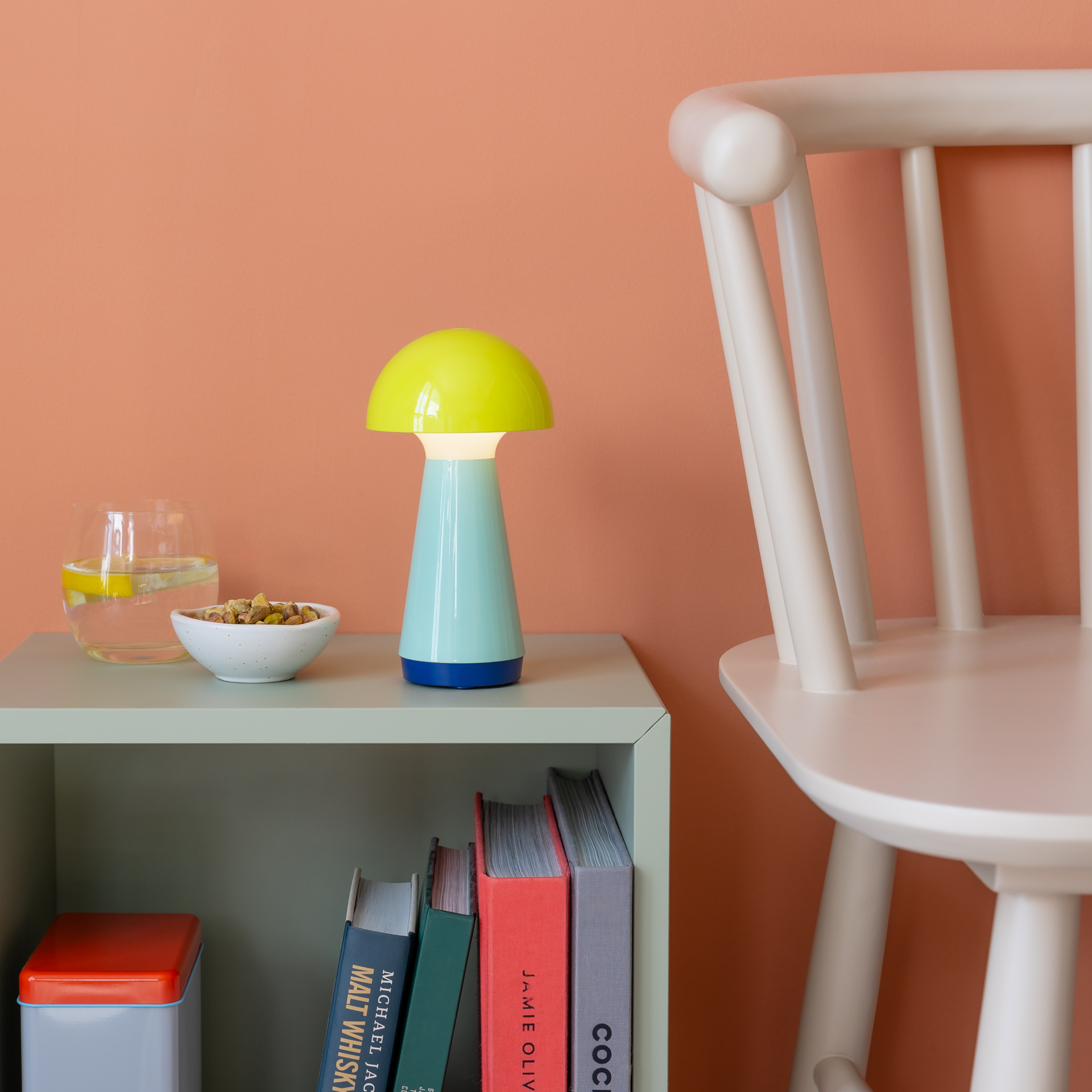 Eine gelbe Tischleuchte Bobbi auf einem Regal neben einem Glas Wasser und einer Schale mit Snacks vor einer orangefarbenen Wand. Ein weißer Stuhl ist im Hintergrund zu sehen.