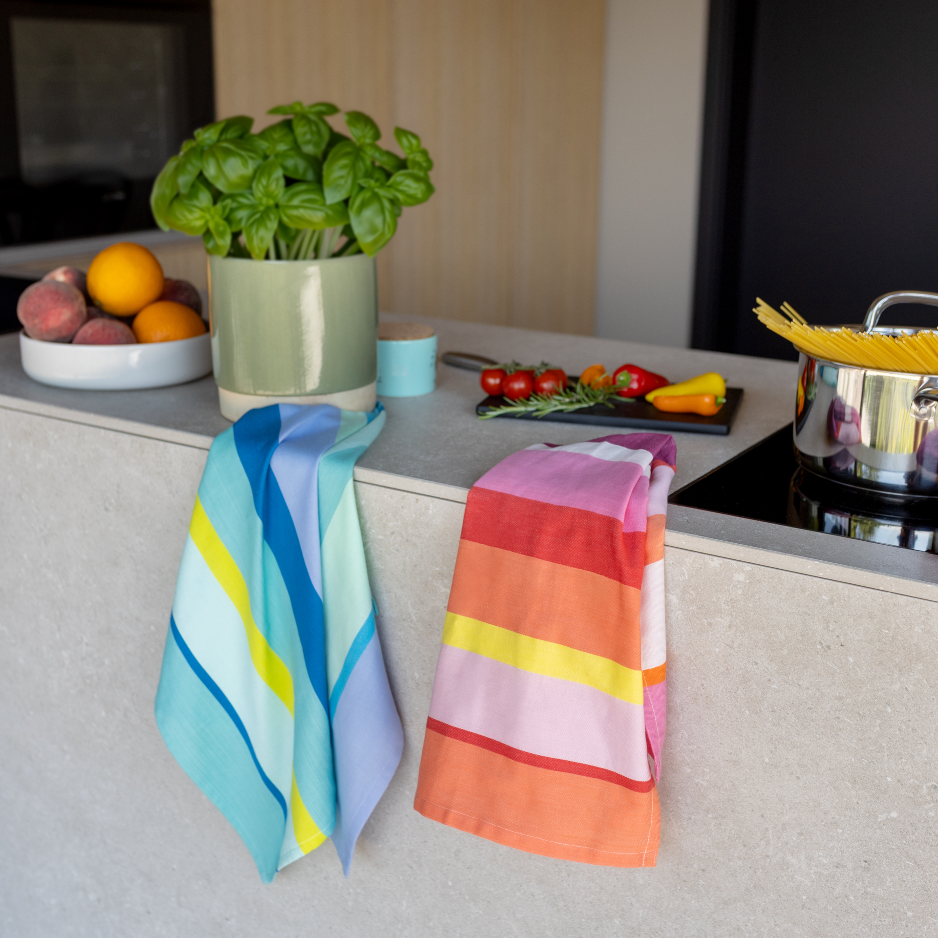 Due coloratissimi canovacci a righe in cotone sono appesi decorativamente sopra un moderno piano di lavoro da cucina. A sinistra blu, turchese, giallo e lilla; a destra rosa, rosso, arancione e giallo. Basilico e frutta adornano la cucina.