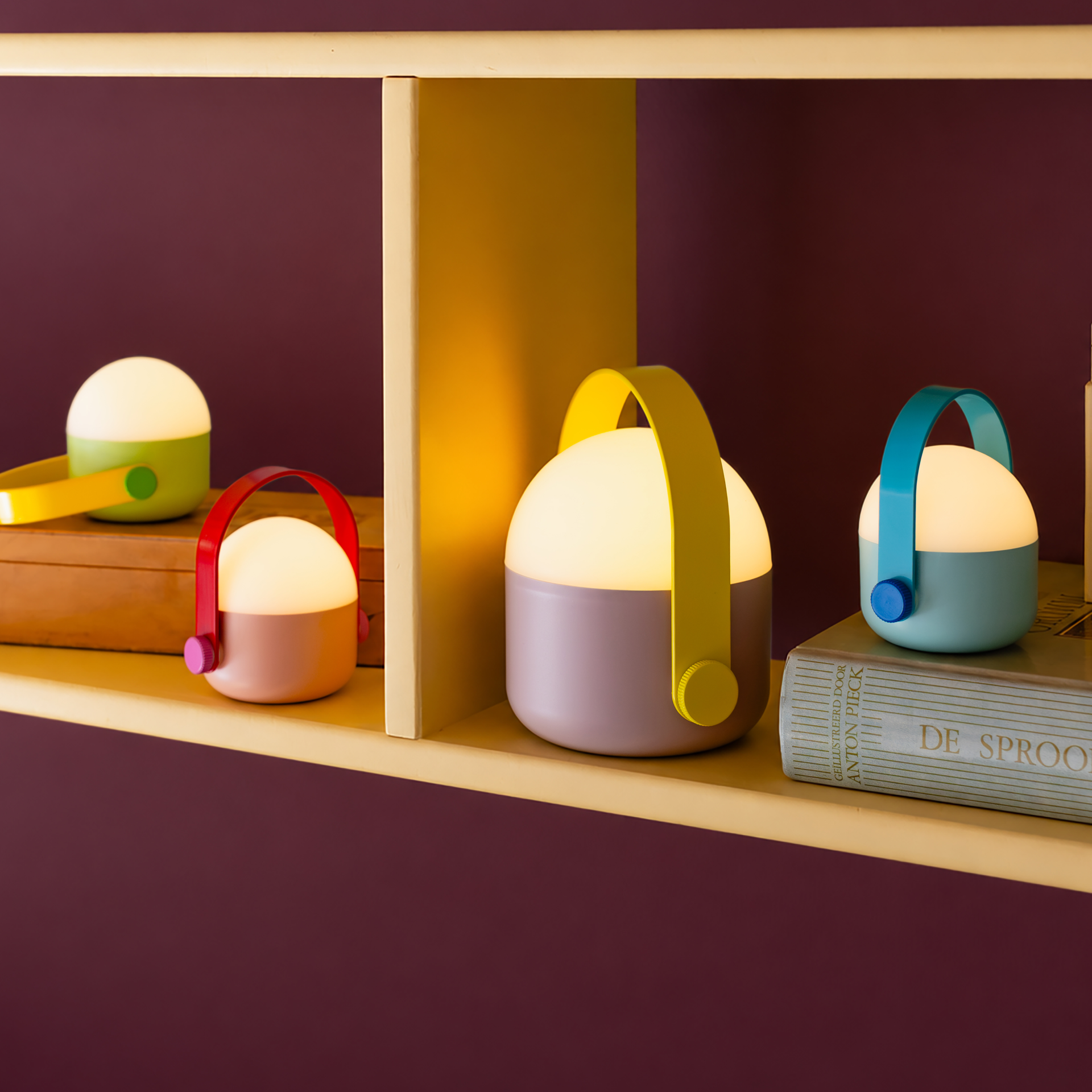 Trois lampes LED à piles aux poignées colorées sont sur une étagère : une verte et jaune sur une boîte en bois, une rose et jaune à côté, et à droite, sur un livre, une bleu clair à poignée turquoise.