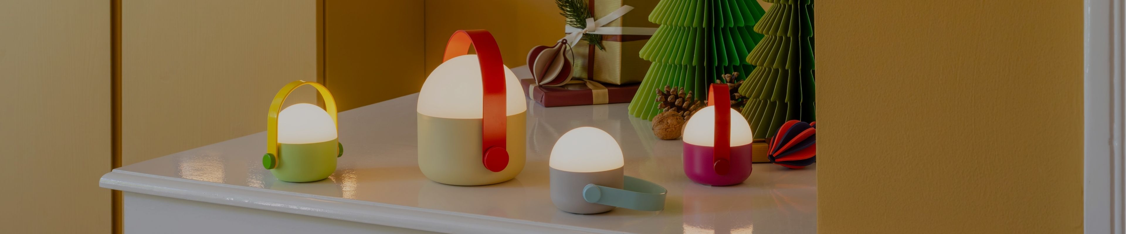 Vier leuchtende, portable Lampen in Limettengrün, Creme, Hellgrau und Magenta stehen auf einer weißen Fläche. Jede mit passendem Griff strahlt warmes Licht. Im Hintergrund: Papierweihnachtsbäume, Geschenke und Tannenzapfen.