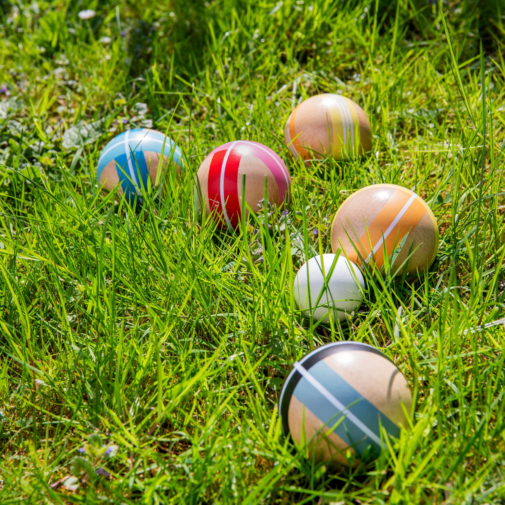 Bunte Holzkugeln für Boule/Boccia liegen als Produktvariante „Set“ verstreut auf sonnigem, hellgrünem Rasen. Die großen Kugeln sind naturfarben mit Streifen in Blau, Rot, Orange und Dunkelgrau. Eine kleine weiße Zielkugel liegt im Gras.