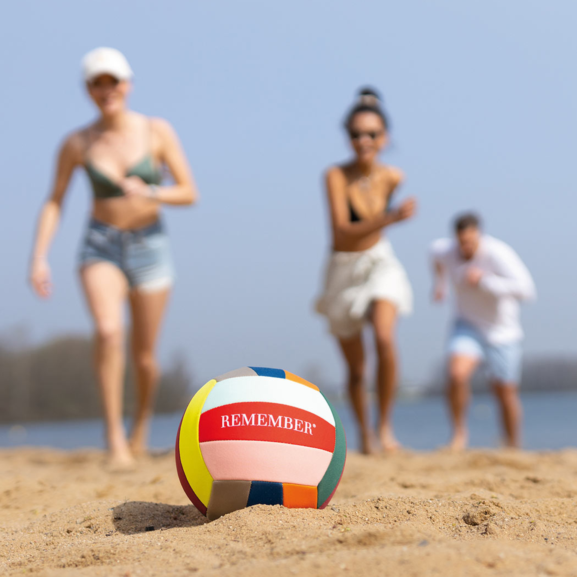 Ein farbiger Freizeit-Ball liegt auf Sand. Im Hintergrund sind drei Personen unscharf zu sehen. Der Ball trägt den Schriftzug "REMEMBER".