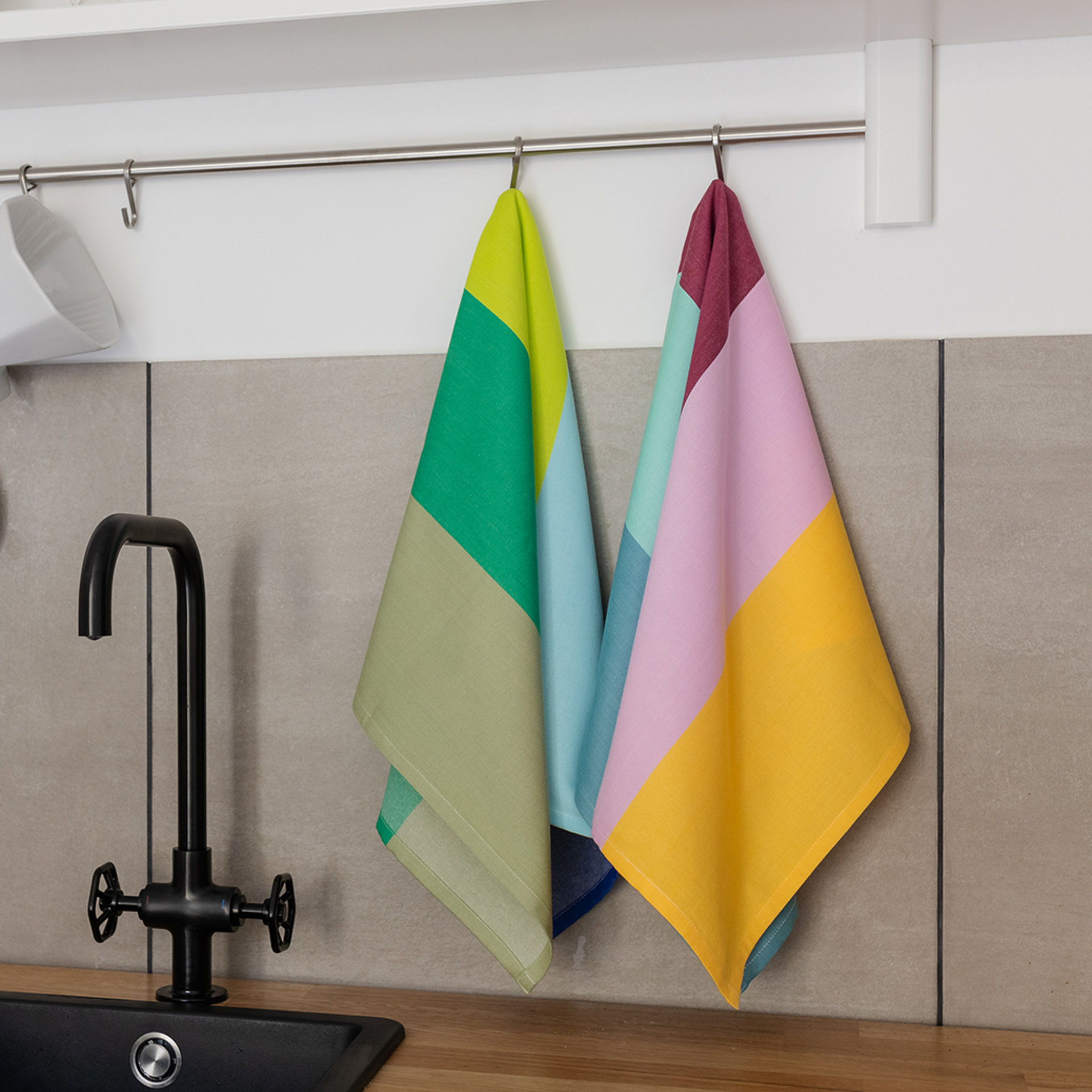 Zwei Trockentücher-Set mit geometrischen Mustern hängen an einem Haken vor einer Küchenspüle. Das linke Tuch ist in Grün- und Blautönen gehalten, das rechte in Gelb- und Rosatönen.