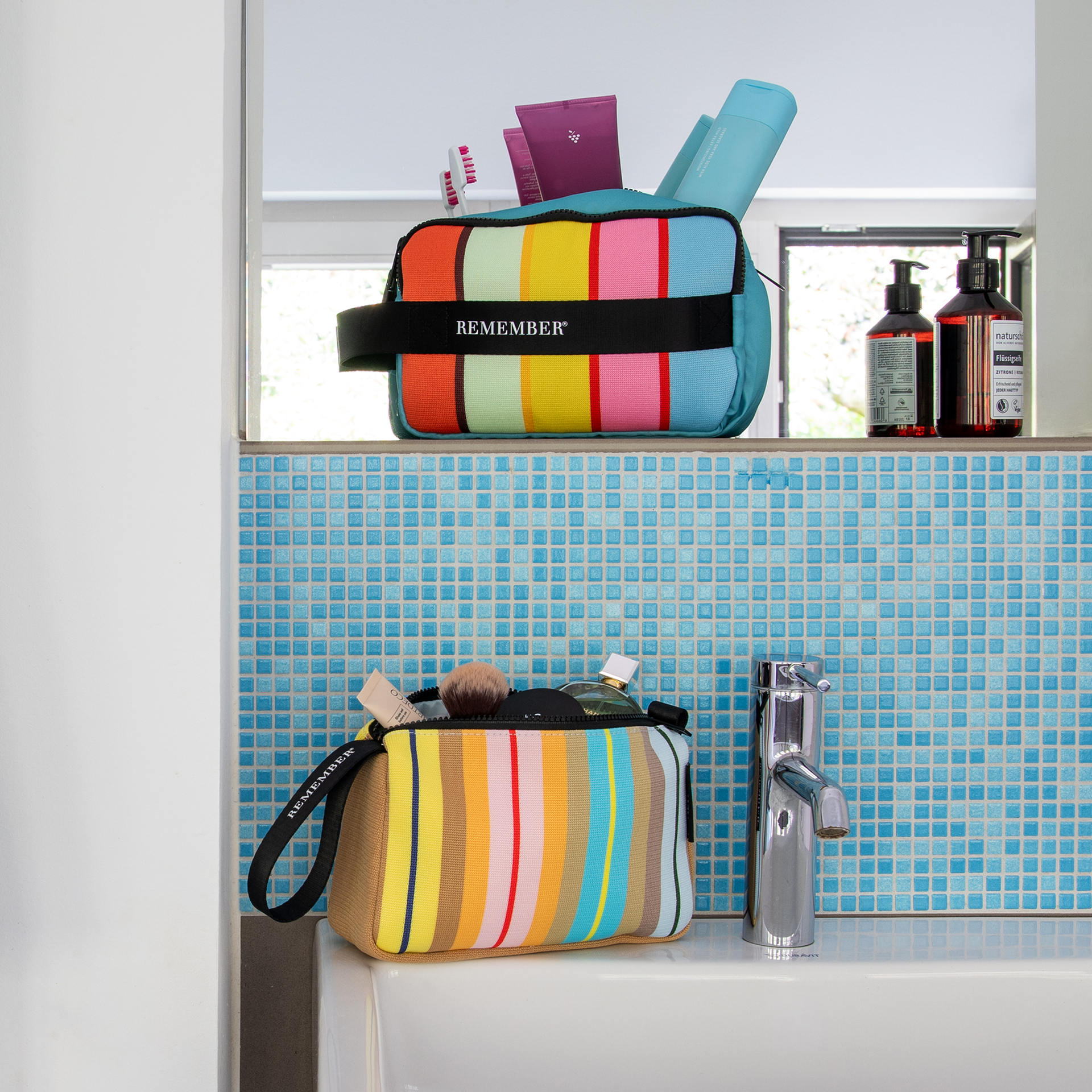 Trousse de toilette rectangulaire, dessus et dessous turquoise. Face avant à larges rayures verticales rouges, vert clair, jaunes, roses et bleu clair. Ruban noir «REMEMBER», fermeture à glissière noire, poignée de transport.