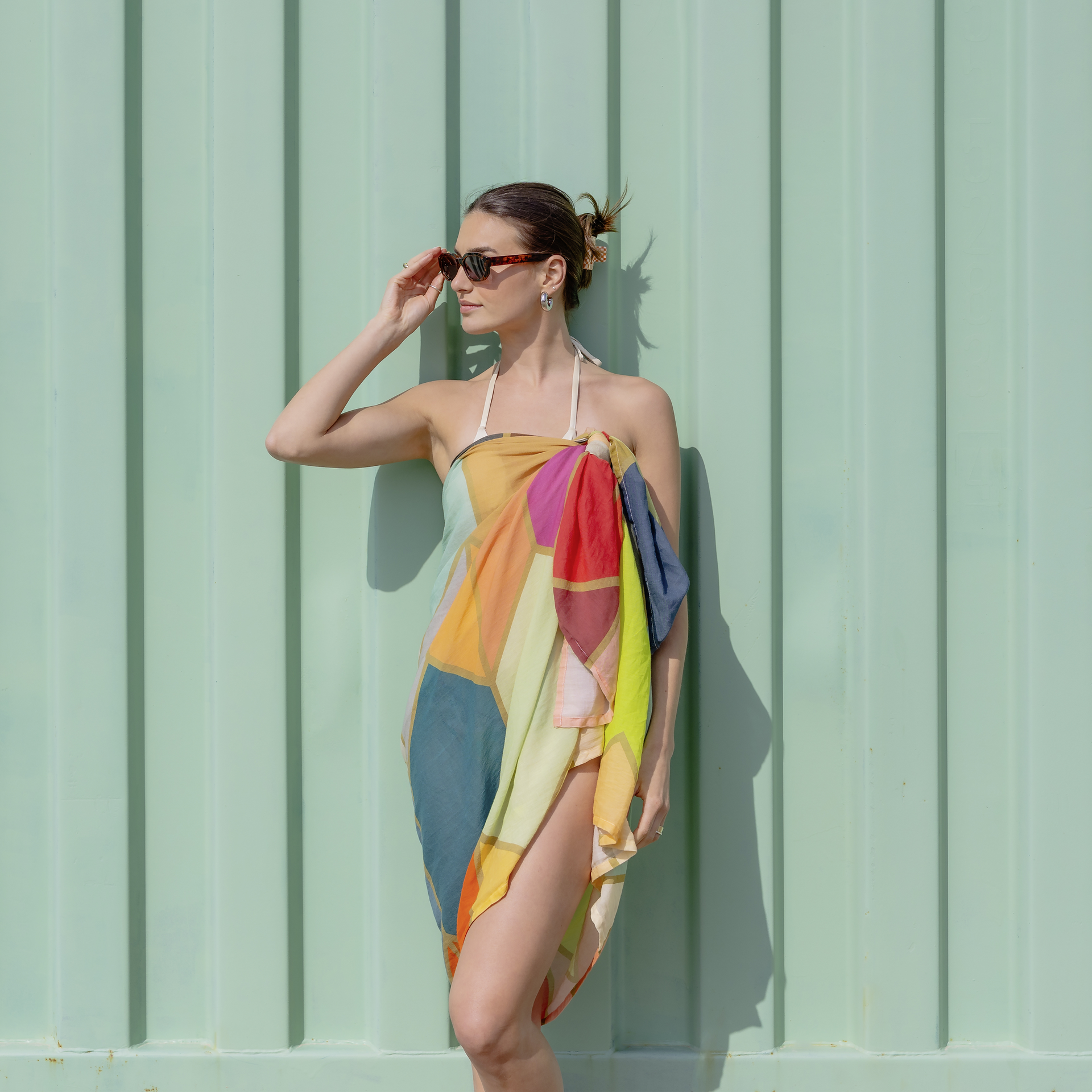 Frau mit Sonnenbrille, die einen bunten Waikiki-Pareo als Kleid vor einer mintgrünen Wand trägt.