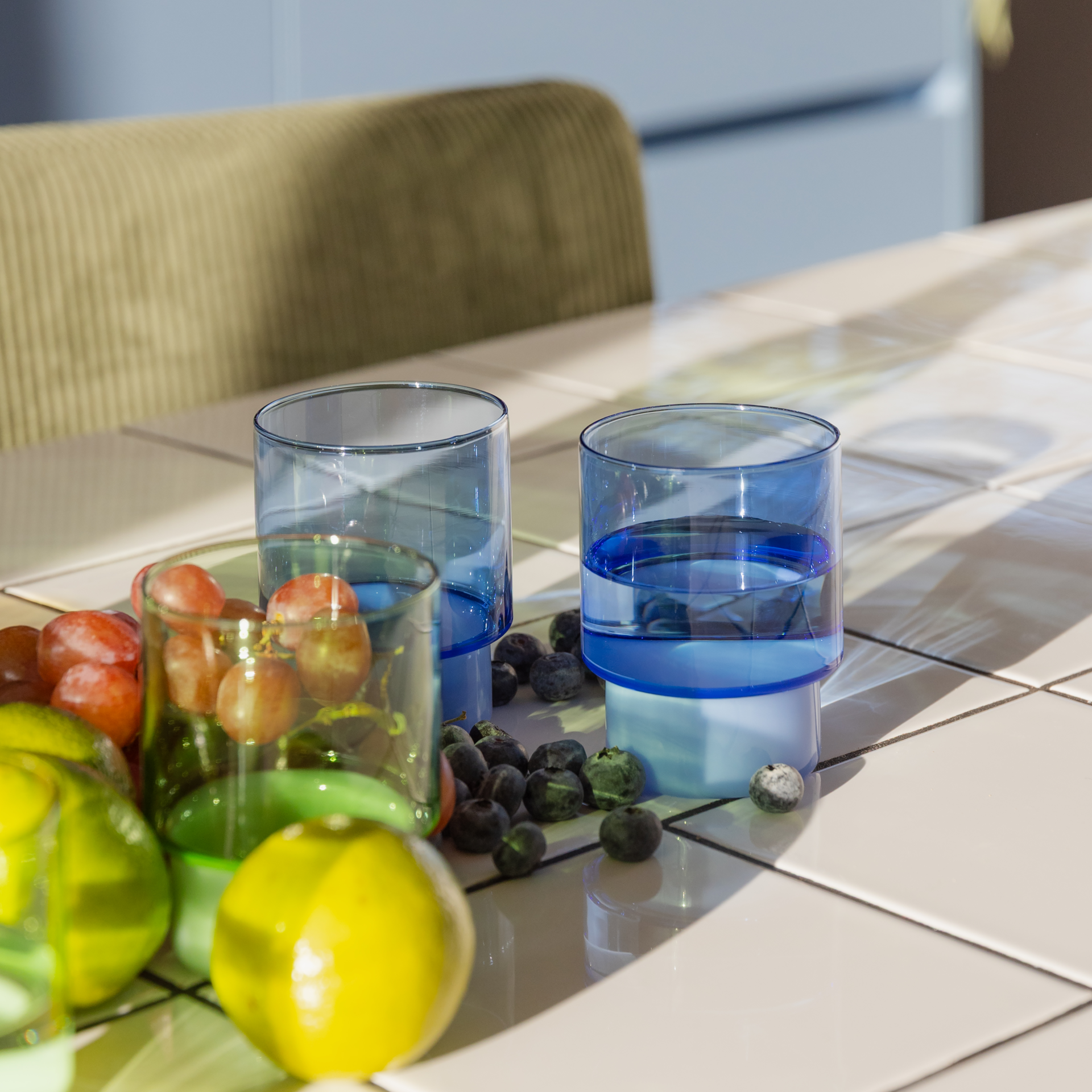 Zwei blaue Gläser, davon eins mit Wasser, sind im Warenkorb und stehen auf einem Tisch. Um sie herum liegen Blaubeeren und weitere Früchte in einem grünen Glas. Der Tisch ist hell und mit Fliesen versehen.