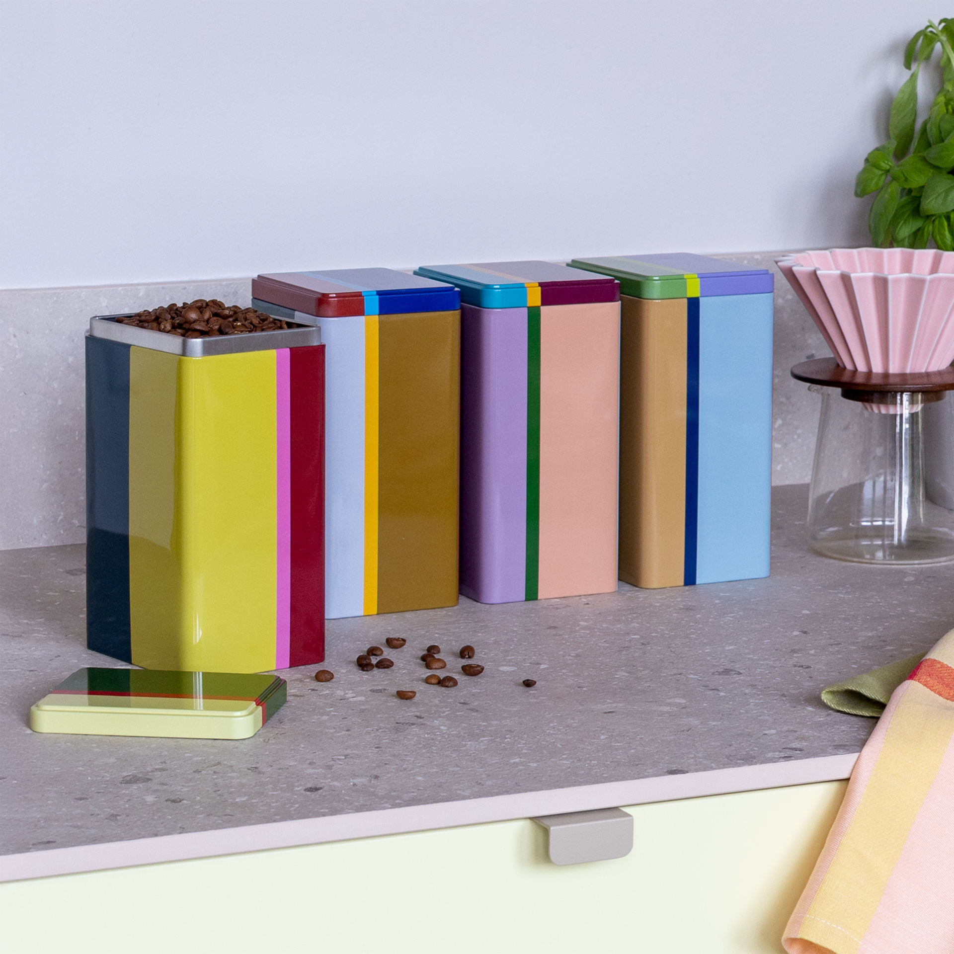 Set di quattro barattoli "Yara" colorati, rettangolari, su un piano di lavoro. Un barattolo è aperto e pieno di chicchi di caffè, con chicchi di caffè sparsi accanto.