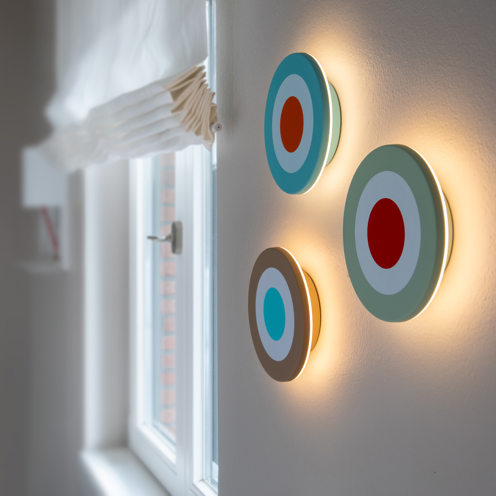 Applique murale circulaire au design minimaliste, composée d’anneaux concentriques : anneau extérieur beige, anneau central blanc et cercle intérieur bleu vif faisant office de source lumineuse. Légèrement inclinée.