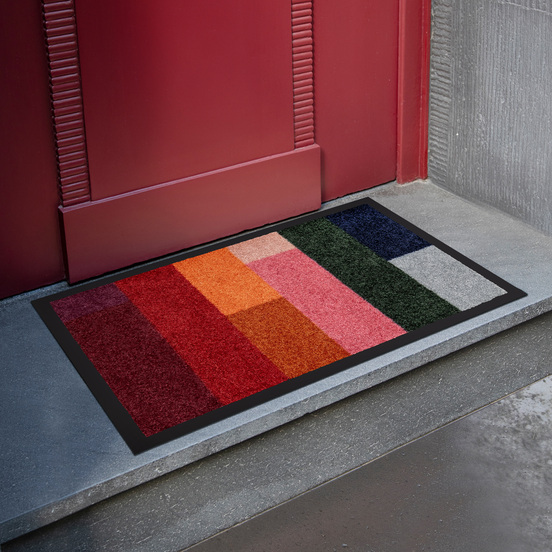 Ein rechteckiges, buntes Fußmatten-Produkt „Nero“ mit Farbverlauf vor einer roten Haustür auf einem grauen Steinboden. Das Fußmatten-Produkt ist in Segmente unterschiedlicher Farben unterteilt.