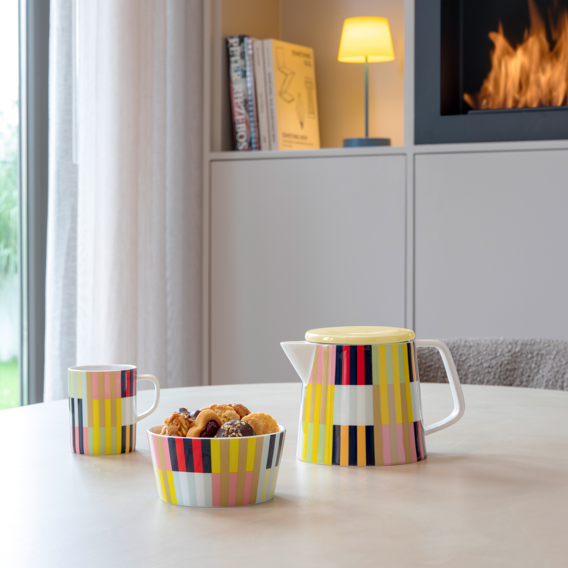 Eine 'Havanna' Teekanne, eine Schale mit Gebäck und eine Tasse, alle mit einem bunten Streifenmuster, stehen auf einem Tisch. Im Hintergrund sind ein Kamin und eine Lampe zu sehen.