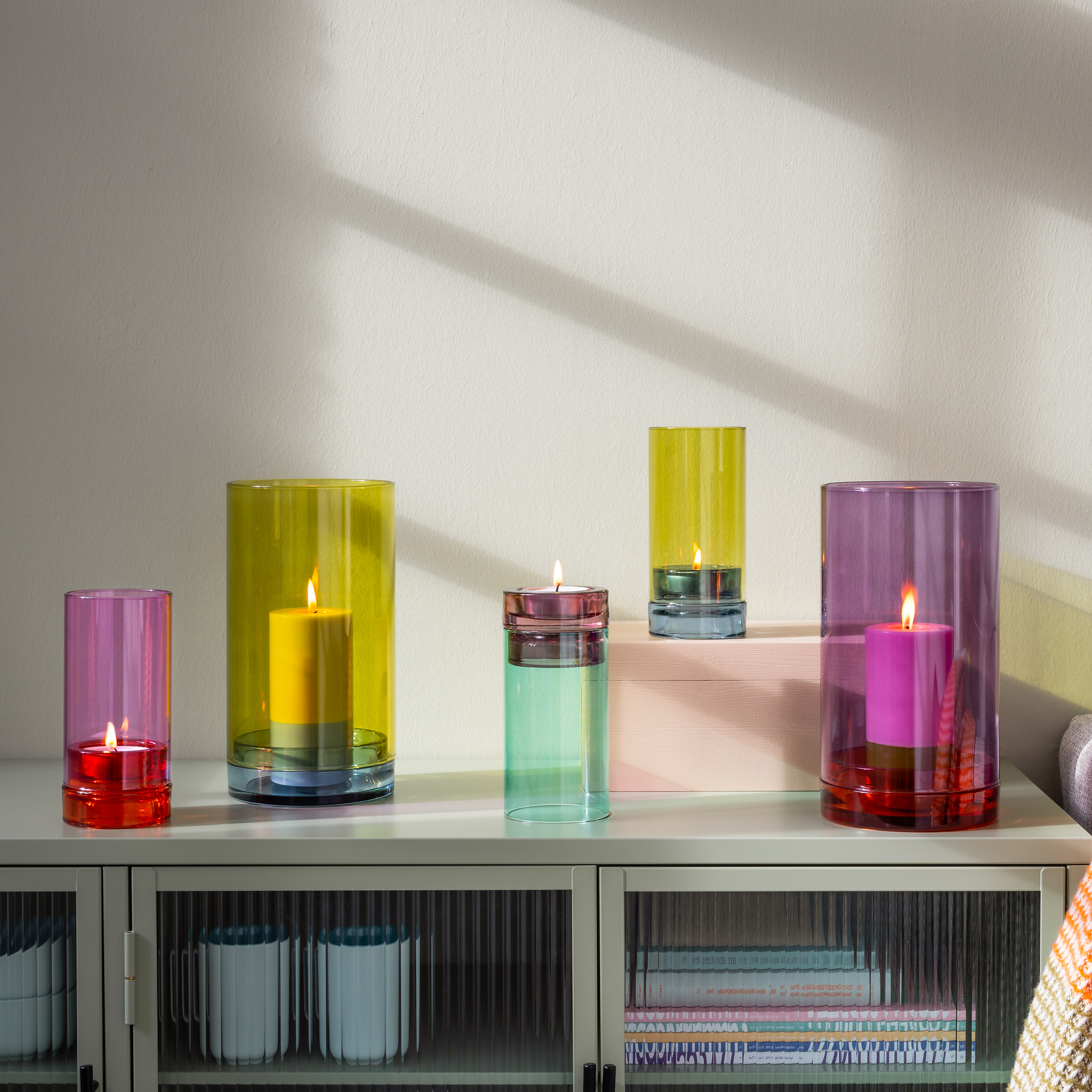 Fünf farbige Glas-Windlichter „Lys“ in Lila, Gelb und Grün mit brennenden Kerzen auf einem Sideboard.