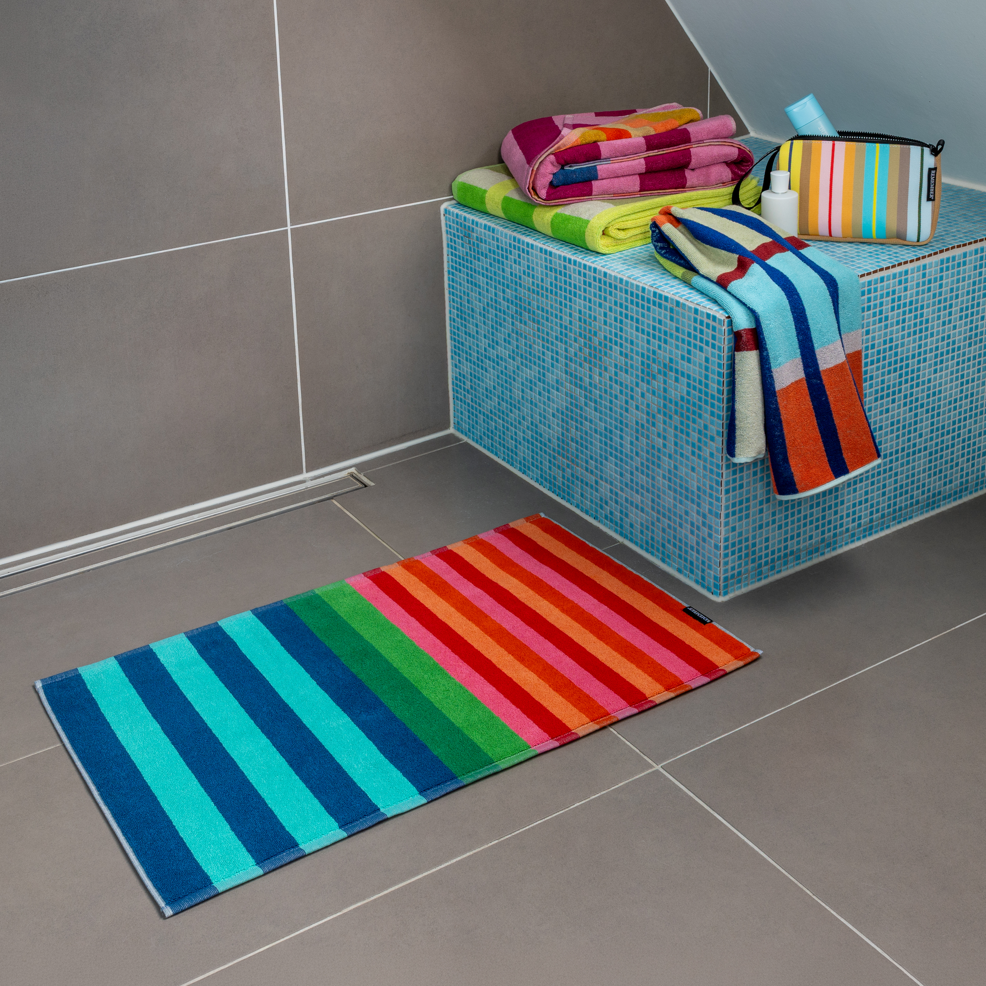 Un tapis de salle de bains Milo, coloré et rayé, est posé sur le carrelage gris. Larges rayures bleu vif, turquoise, vert, rouge, orange et rose. À l'arrière-plan, serviettes pliées et trousse assortie.
