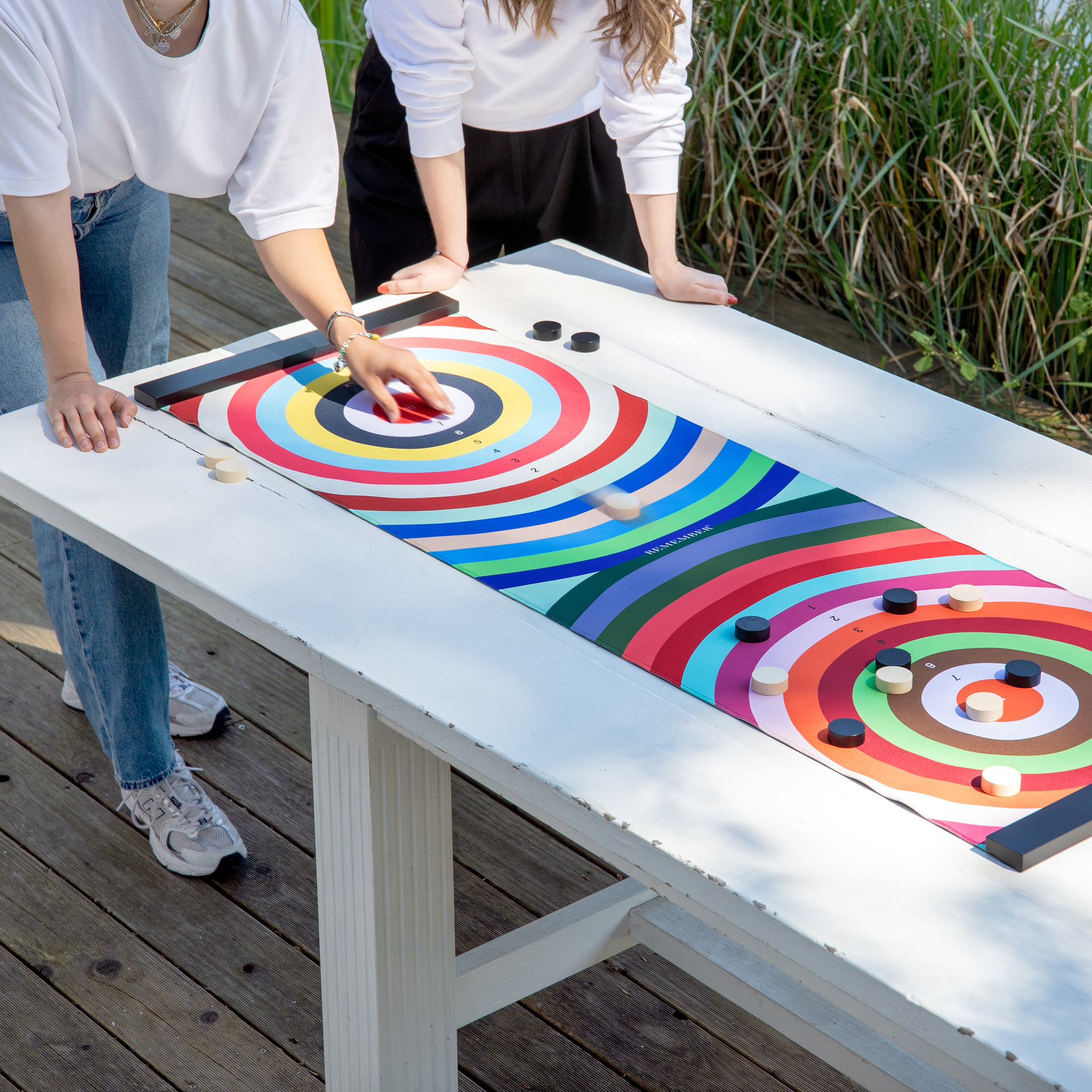 Zwei Personen spielen draußen ein buntes Brettspiel auf einem weißen Tisch. Eine Hand schiebt eine beige Figur; schwarze und beige Pucks liegen auf nummerierten Feldern.