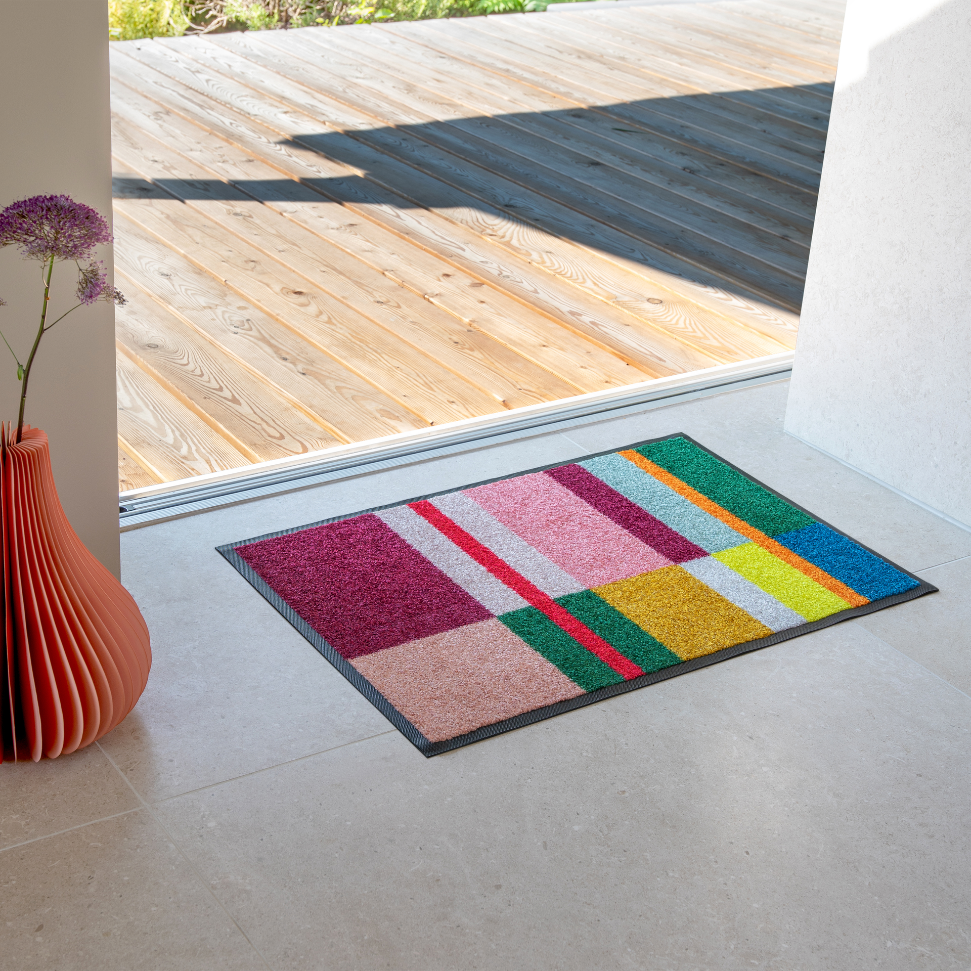 Ein farbenfroher Fußabtreter mit geometrischem Muster in Magenta, Rosa, Rot, Grün, Gelb, Blau und Beige liegt auf hellgrauen Fliesen. Links steht eine geriffelte orange Vase mit violetter Alliumblüte.