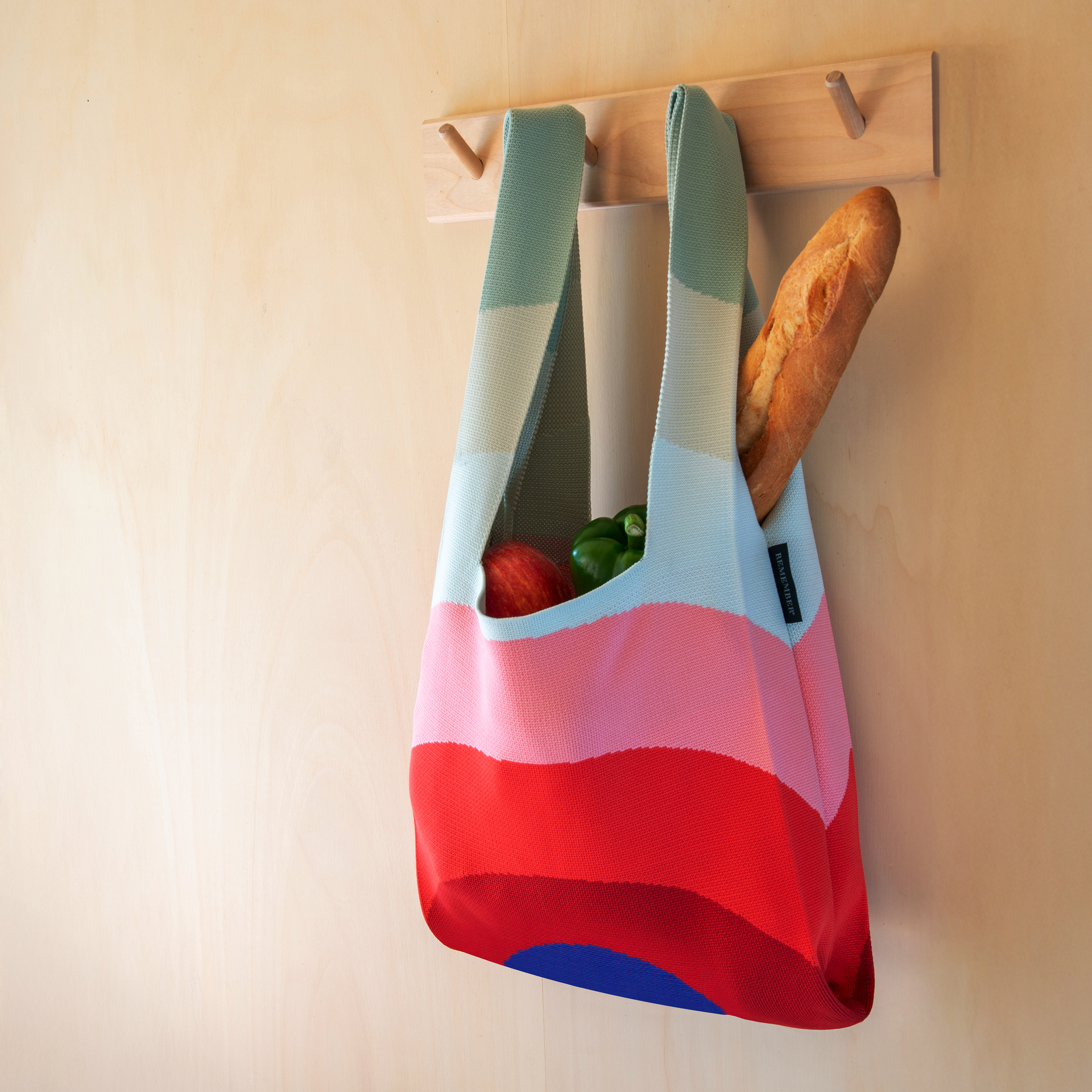 Das Produkt „Gehäkelte Stricktasche 'Indigo'“ wird als Lifestyle-Accessoire am Point of Sale präsentiert; im Lieferumfang enthalten sind Brot, Apfel und Paprika. Das Design umfasst bunte Streifen.
