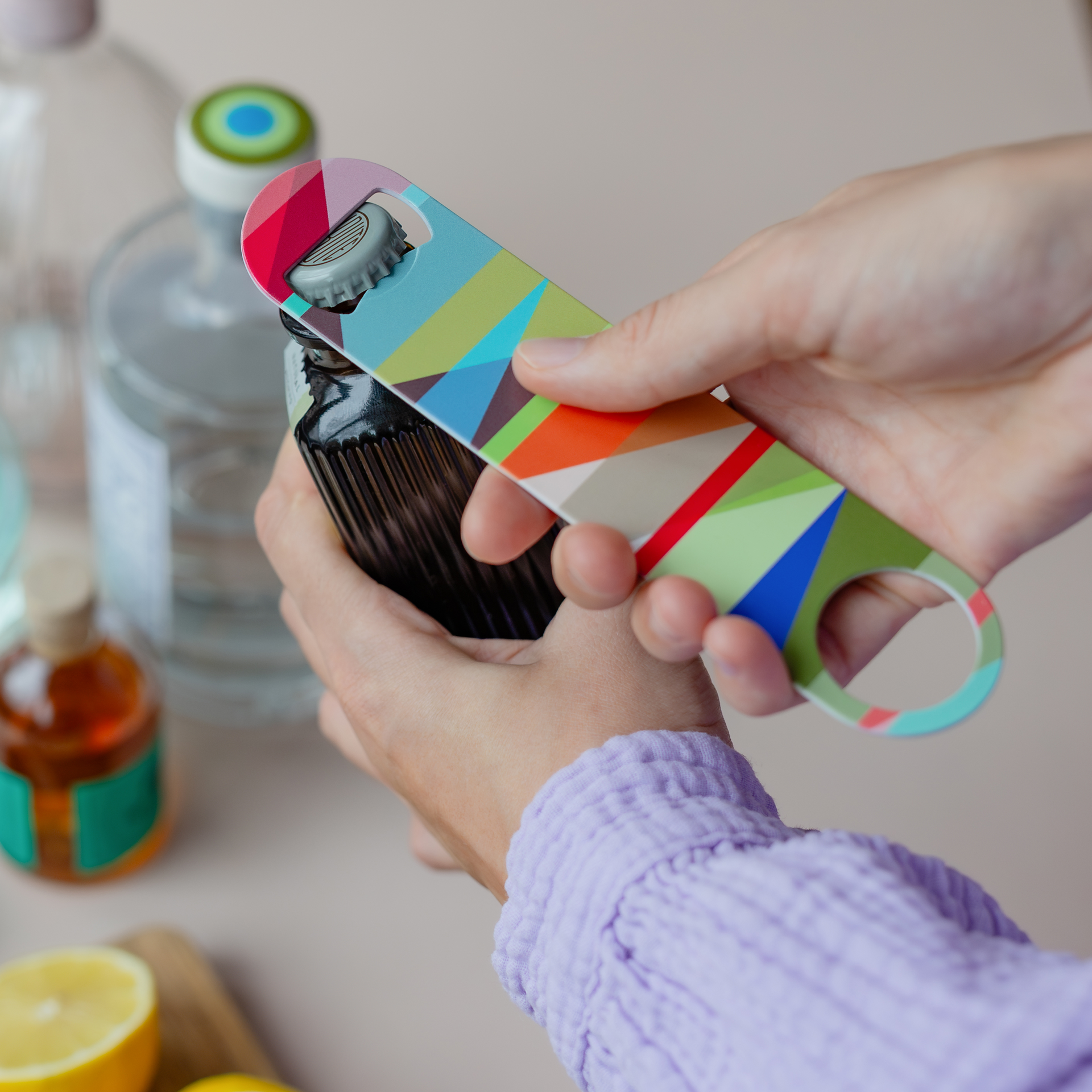 Hände öffnen mit dem bunten 'Mateo' Flaschenöffner eine dunkle Flasche. Im Hintergrund sind weitere Flaschen erkennbar.