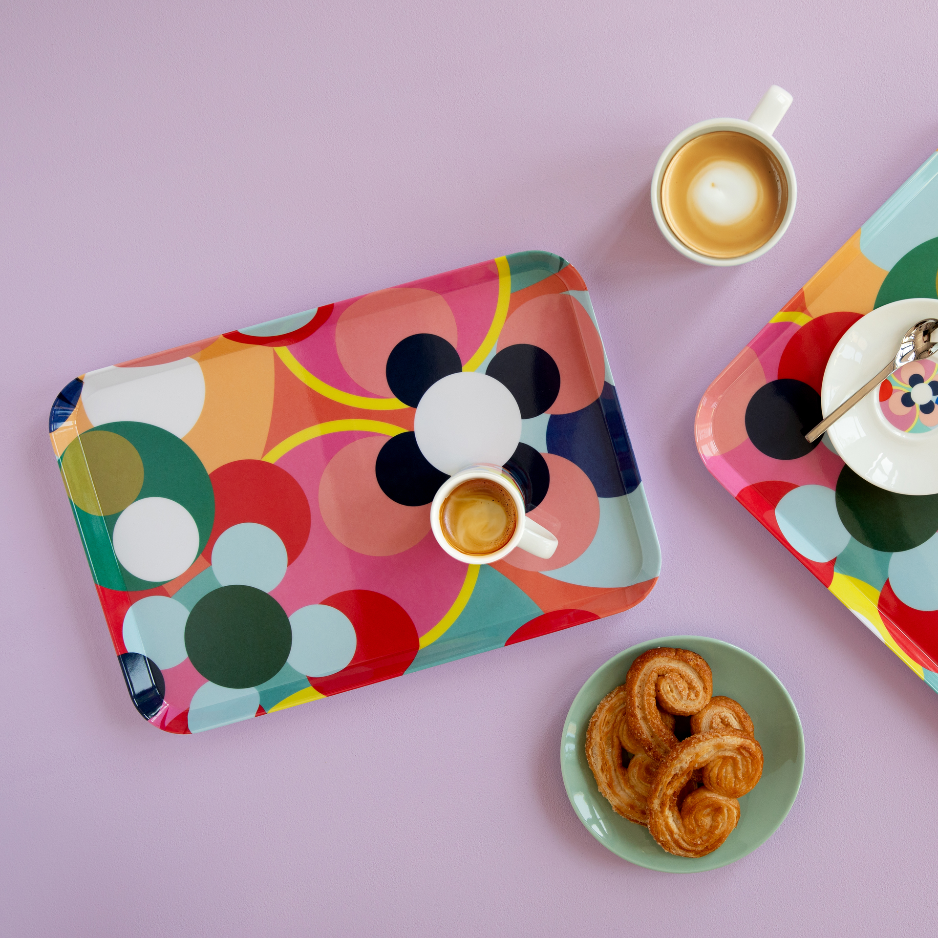 Zwei Mini Tabletts mit Muster, Kaffee und Gebäck auf lila Untergrund. Das Tablett links hat ein Blumenmuster und trägt eine Espressotasse.