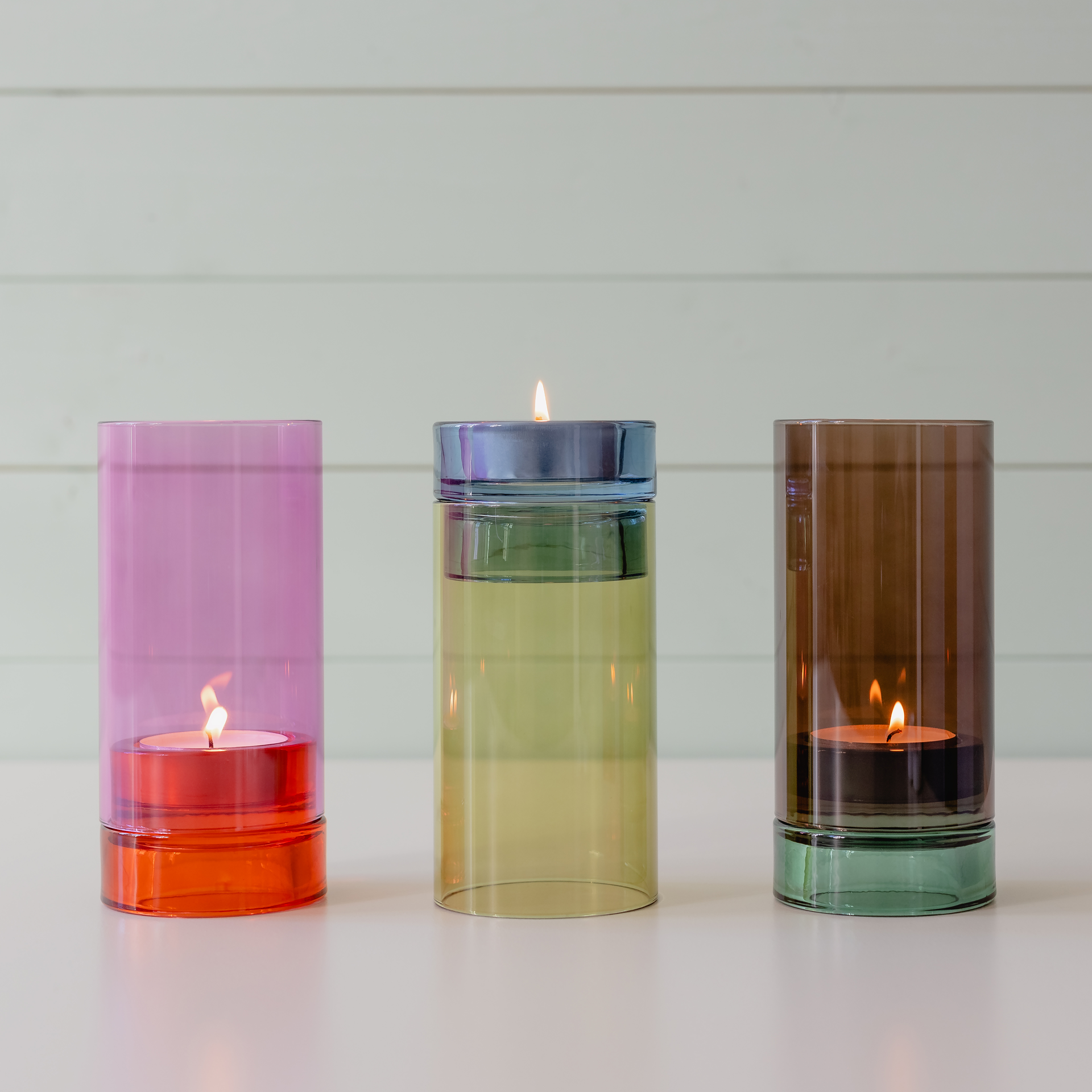 Drei farbige Windlichter 'Lys' aus Glas mit Teelichtern stehen nebeneinander. Das linke ist pink, das mittlere gelbgrün und das rechte braun.