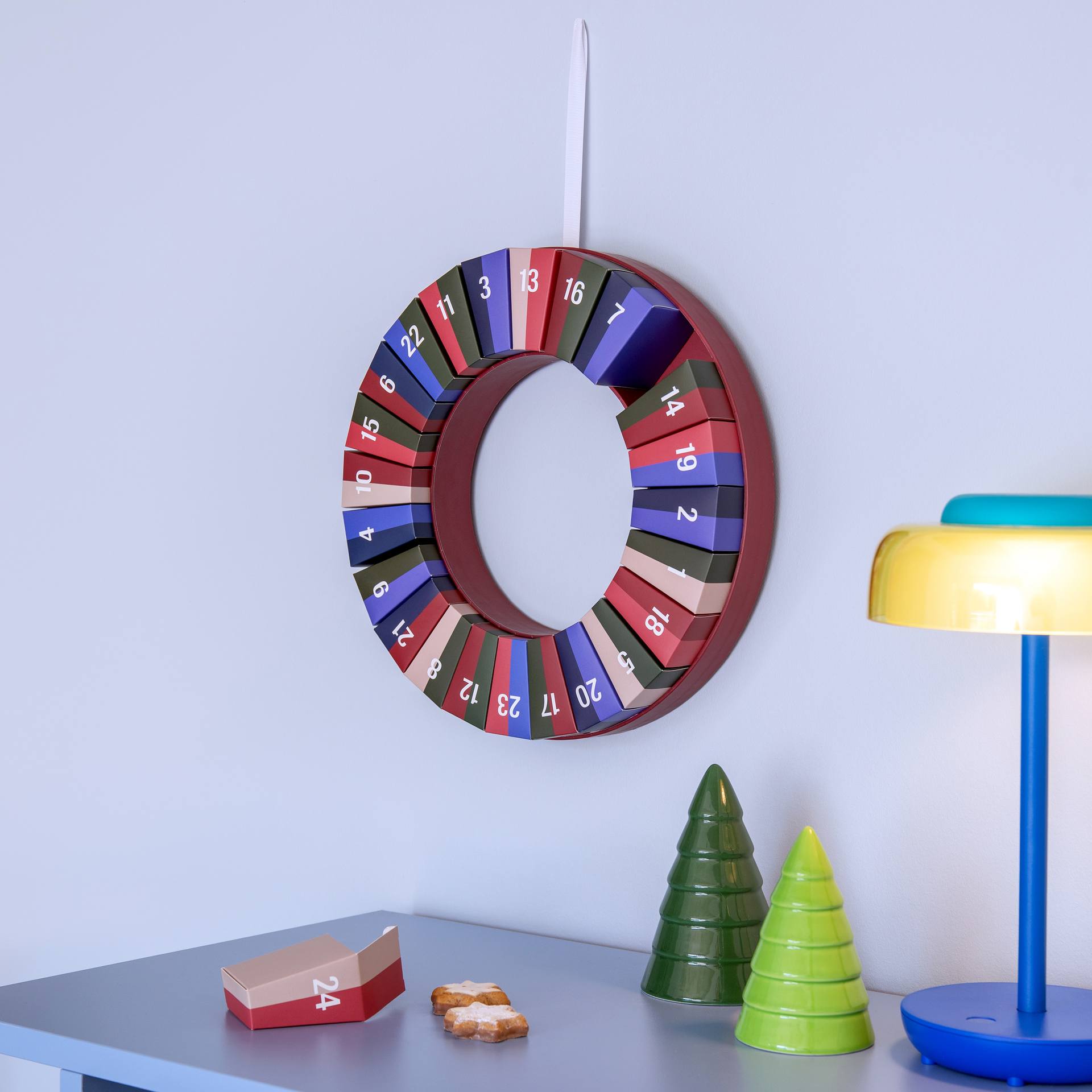 Couronne de l'Avent ronde avec 24 cases numérotées de différentes couleurs (bleu foncé, vert foncé, rose, beige). Suspendue à un ruban blanc, elle affiche les chiffres de 1 à 24 répartis régulièrement autour du centre.