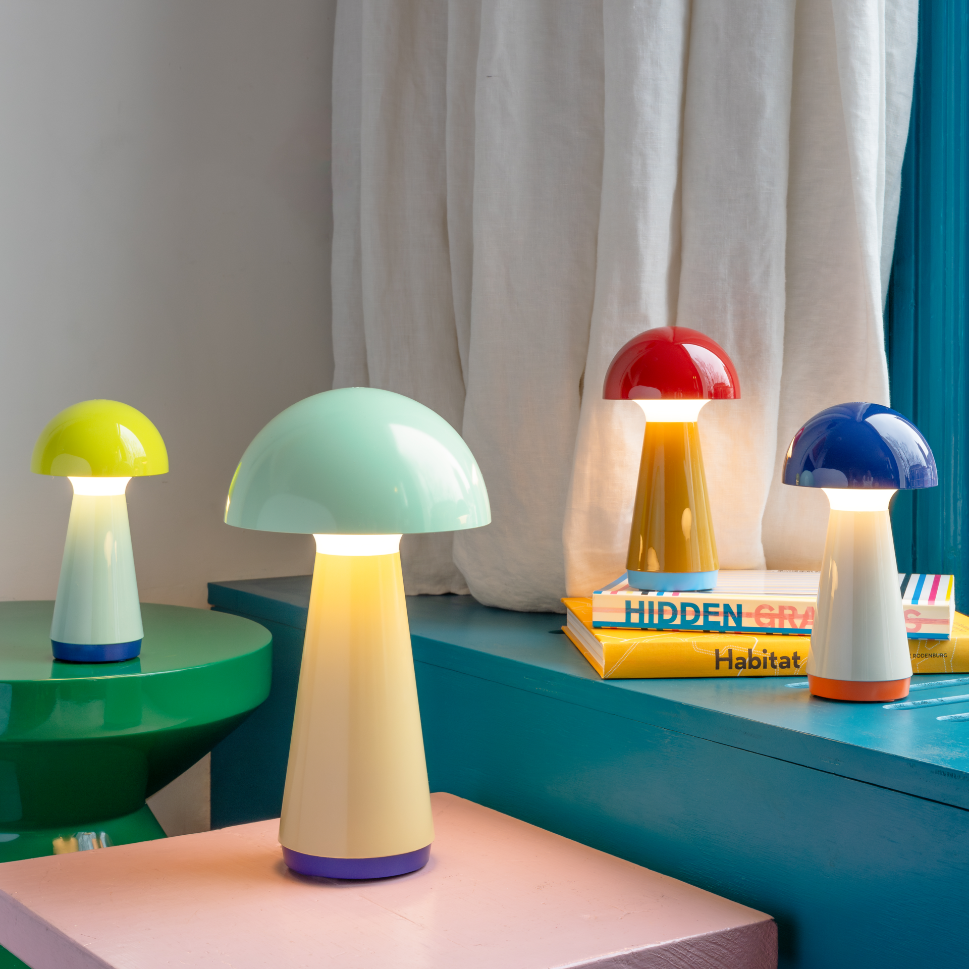 Vier pilzförmige Tischleuchten 'Bob' in Mint, Gelb, Rot und Blau leuchten auf farbigen Tischen und Bücherstapeln vor Vorhängen und einer Wand.