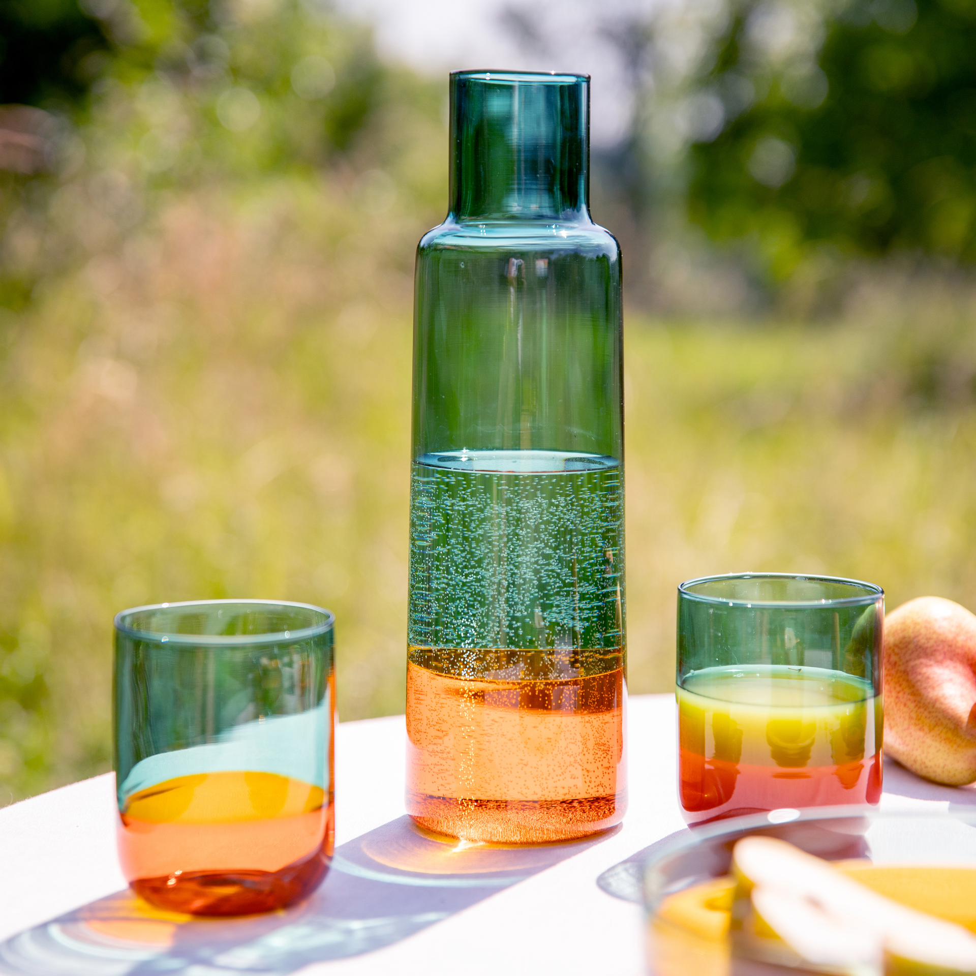 Eine schlanke Glaskaraffe 'amber' gefüllt mit einer sprudelnden Flüssigkeit, flankiert von zwei passenden Gläsern auf einem Tisch im Freien, der Hintergrund ist unscharf.