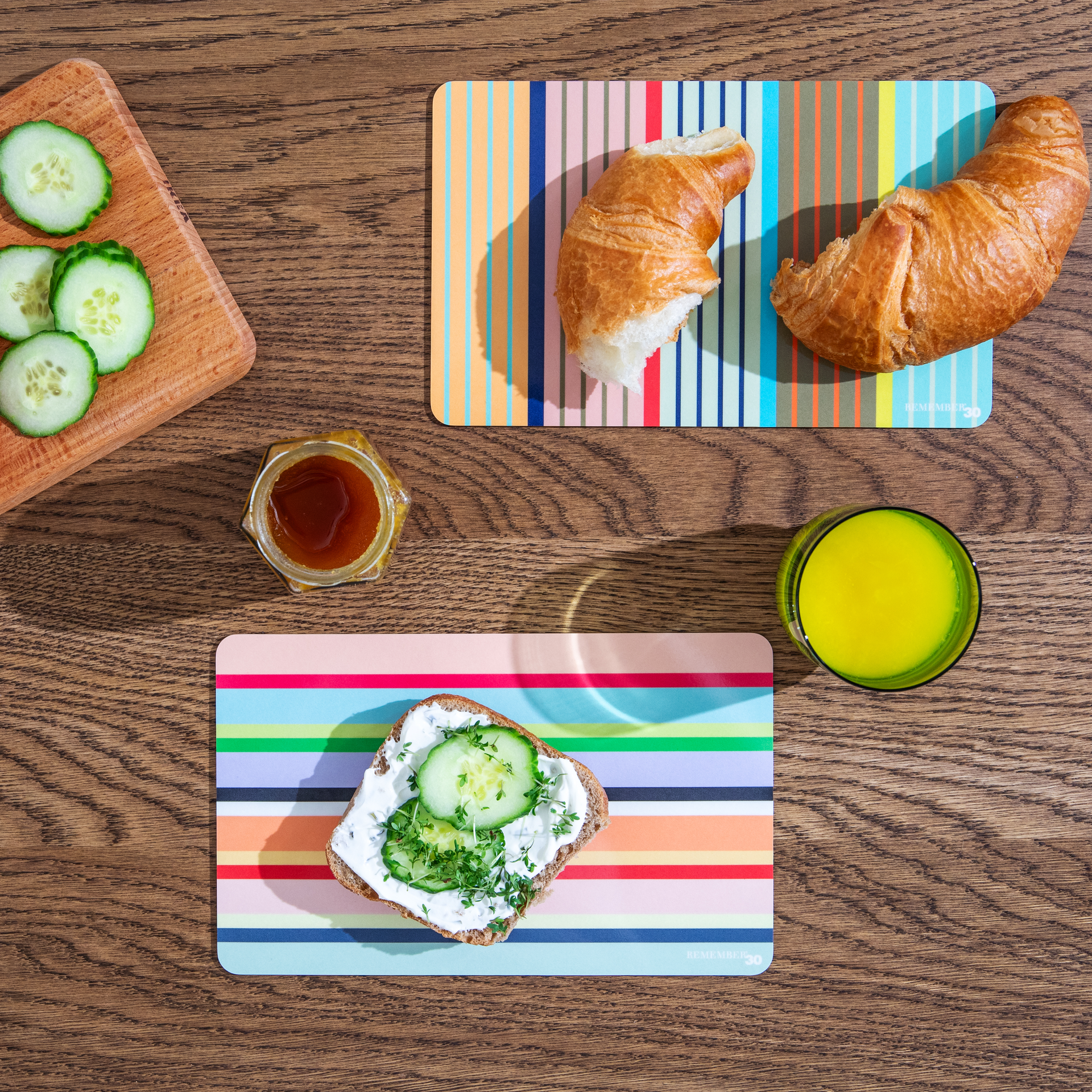Vista dall'alto: tavola da colazione a righe con croissant, pane con crema di formaggio e cetrioli, miele e succo di frutta su un tavolo di legno.