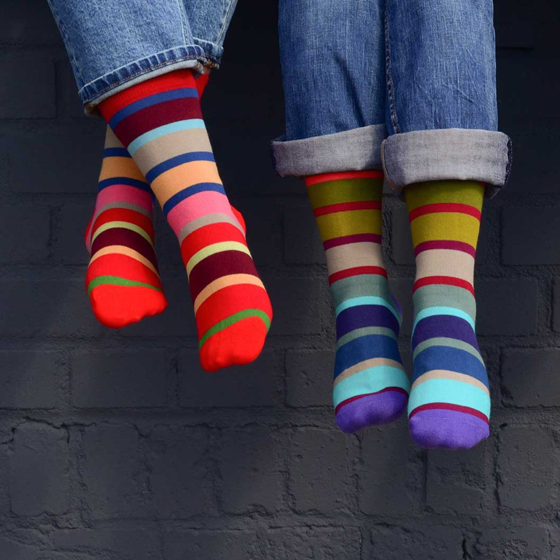 Zwei Personen in Jeans tragen farbenfrohe, gestreifte Socken in Rot- und Blautönen vor einer dunklen Ziegelwand. Modell 33, Gr. 41-46.