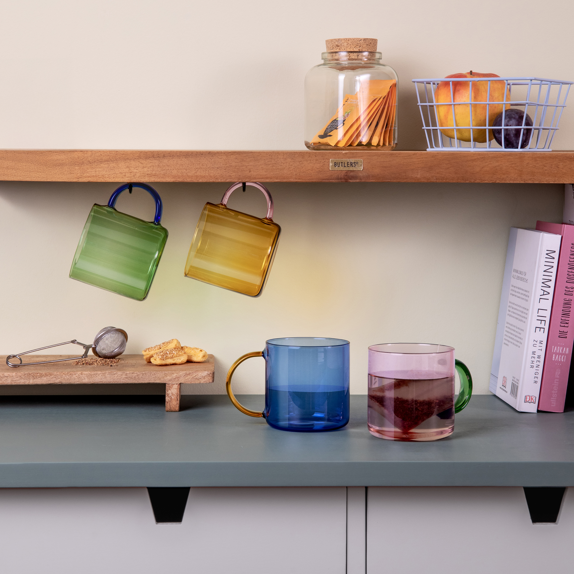 Zwei Glasbecher 'Fred' im Set, einer grün-blau hängend, einer blau stehend; zweiter Becher rosa mit Teebeutel, diverse Küchenutensilien und Deko im Hintergrund.