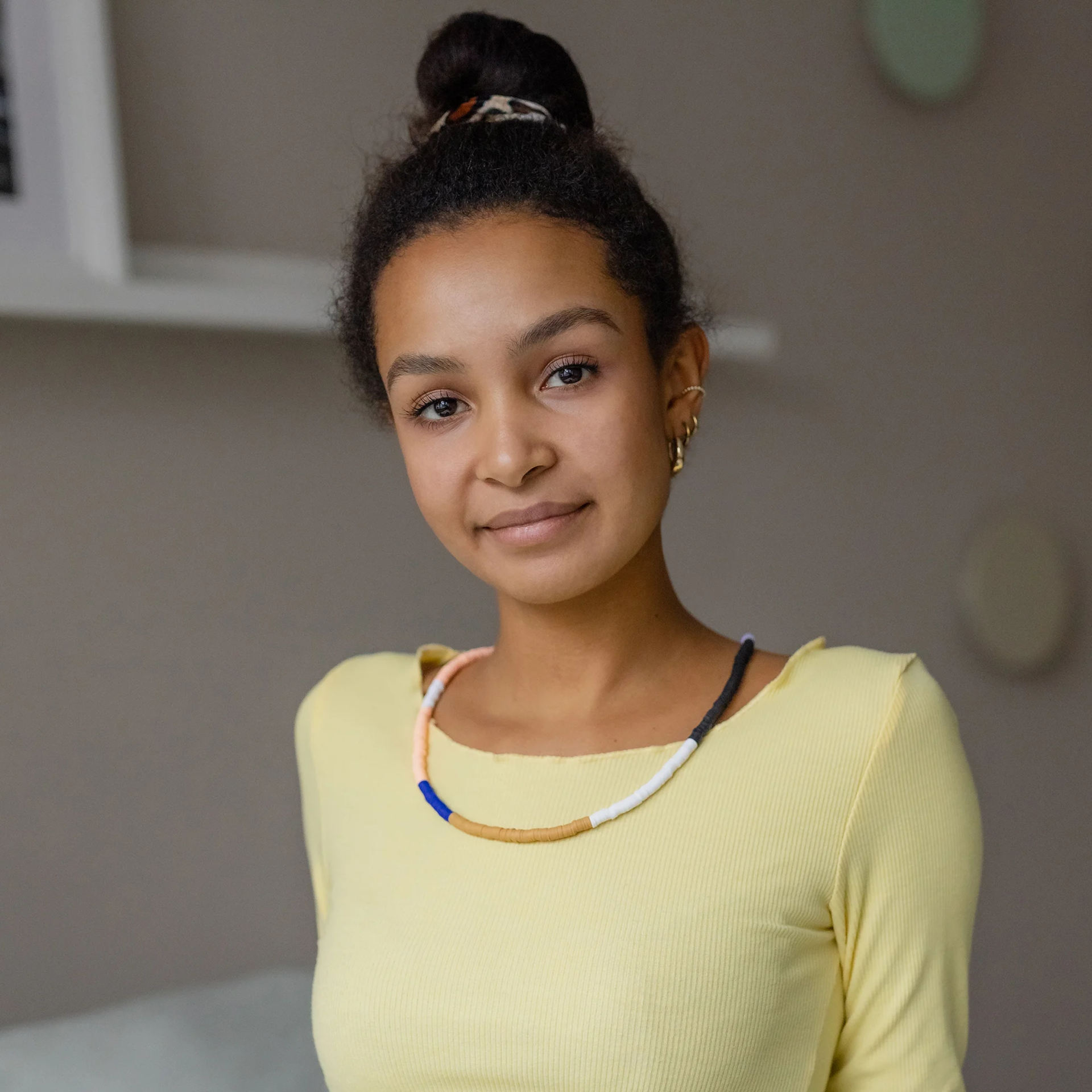 Junge Frau mit dunkler Haut und dunklem, lockigem Haar im Dutt blickt mit sanftem Lächeln in die Kamera. Sie trägt ein geripptes, hellgelbes Langarmshirt, bunte Perlenkette und goldene Ohrringe vor grauem Hintergrund.