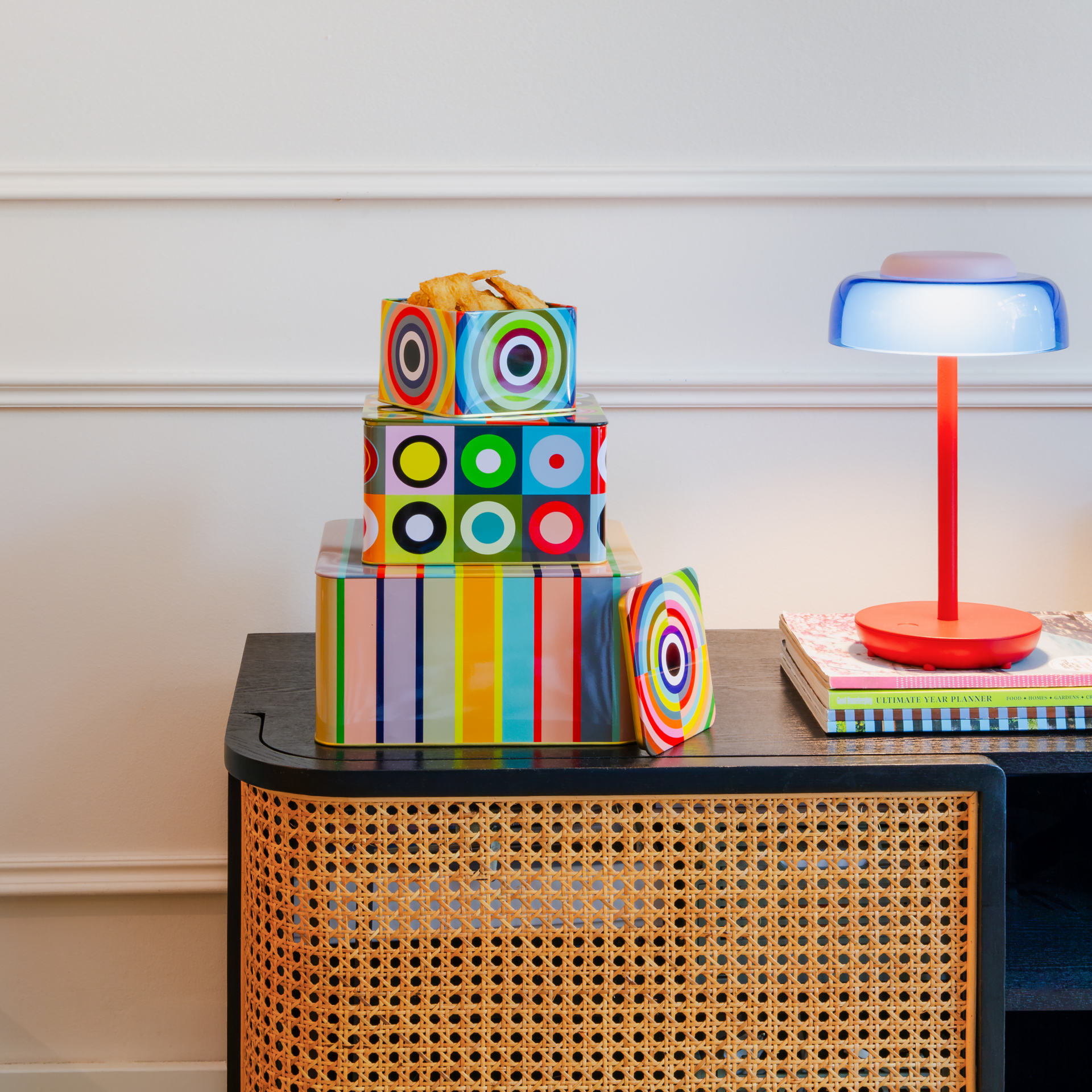 Trois boîtes en fer-blanc décoratives sont empilées. La plus grande a des rayures verticales colorées. Celle du milieu a des carrés et des cercles concentriques multicolores. En haut, la plus petite a un motif similaire. « 30 » à gauche.