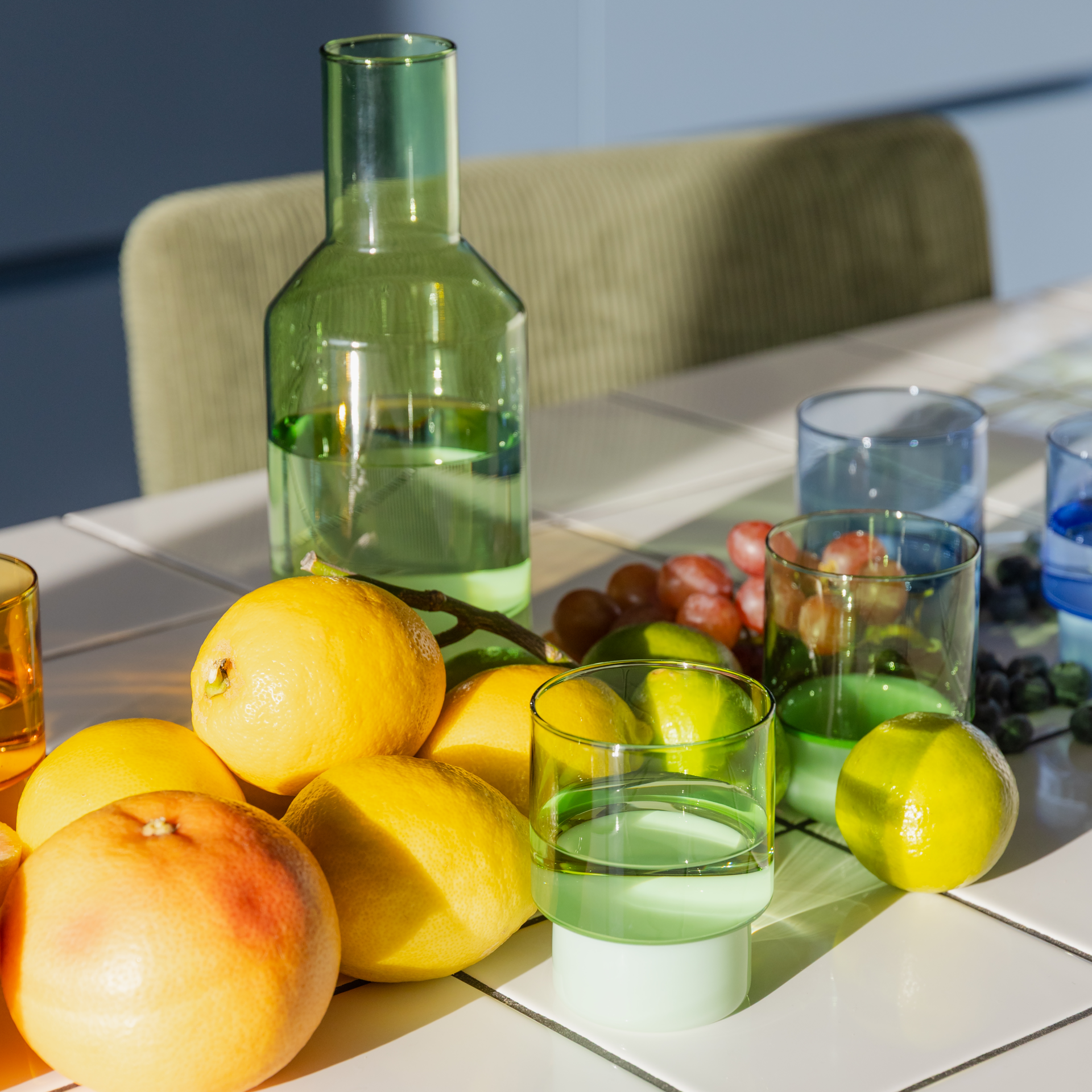 Ein Tisch mit grünem Gläser-Set, Karaffe und Zitrusfrüchten. Daneben liegen blaue Gläser mit Weintrauben und Heidelbeeren.